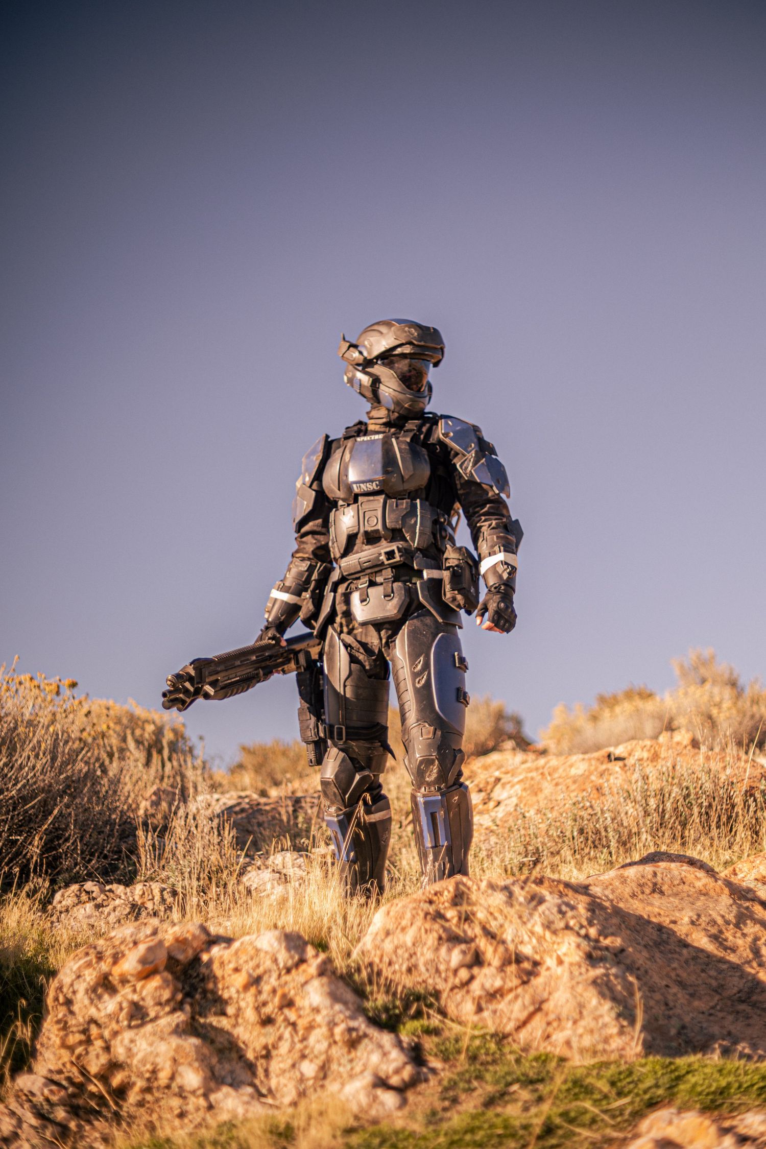 Cosplayer in dark futuristic battle armor carrying a sci-fi weapon while standing on rocky desert terrain.