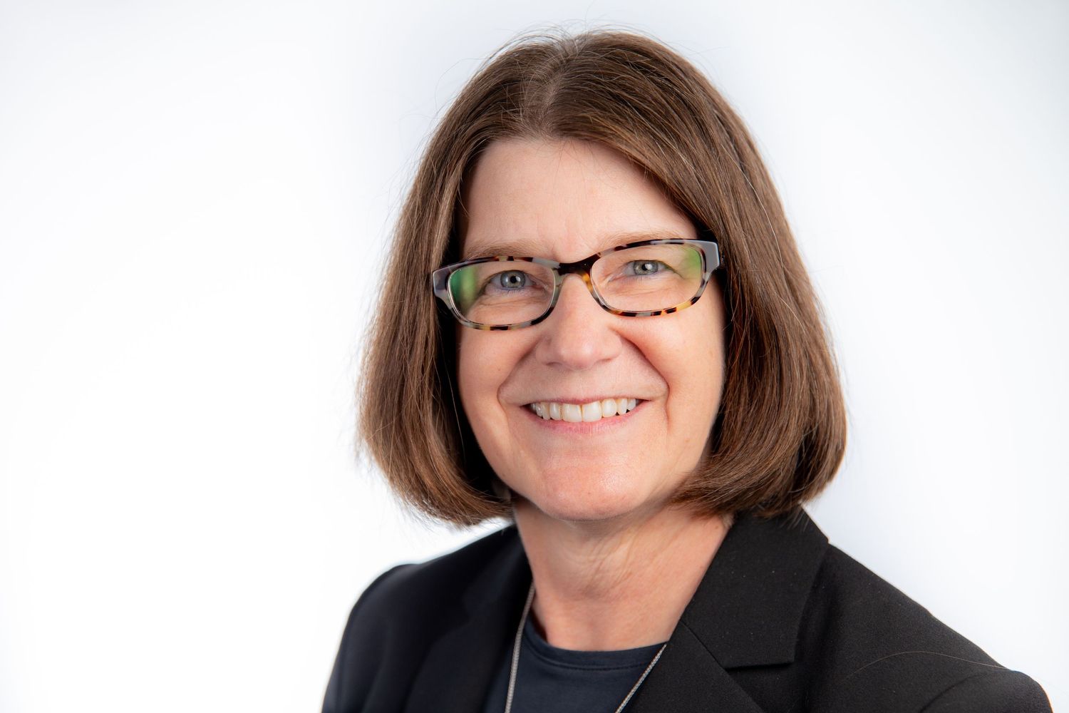 Professional headshot of a smiling woman with short brown hair and glasses wearing a black blazer on white background.