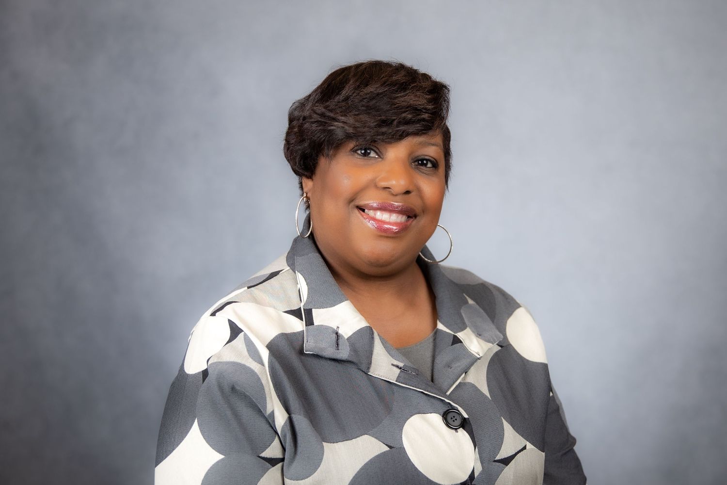 Smiling woman with dark hair wearing gray floral blouse and hoop earrings on gray background.