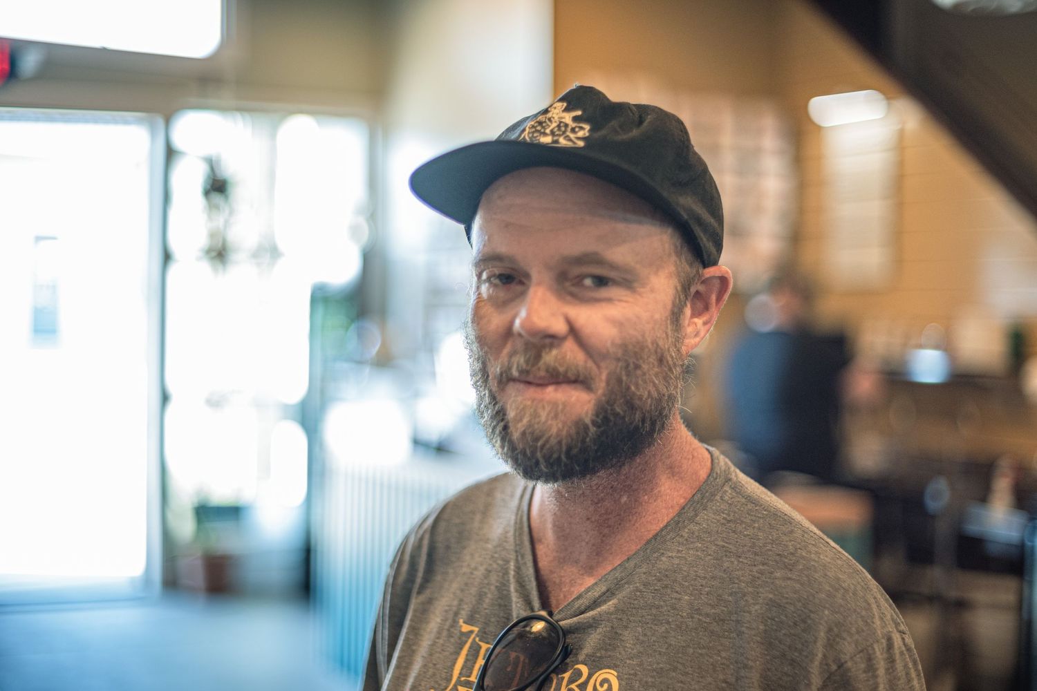 Bearded man in a black cap and gray t-shirt smiling inside a casual restaurant or coffee shop.