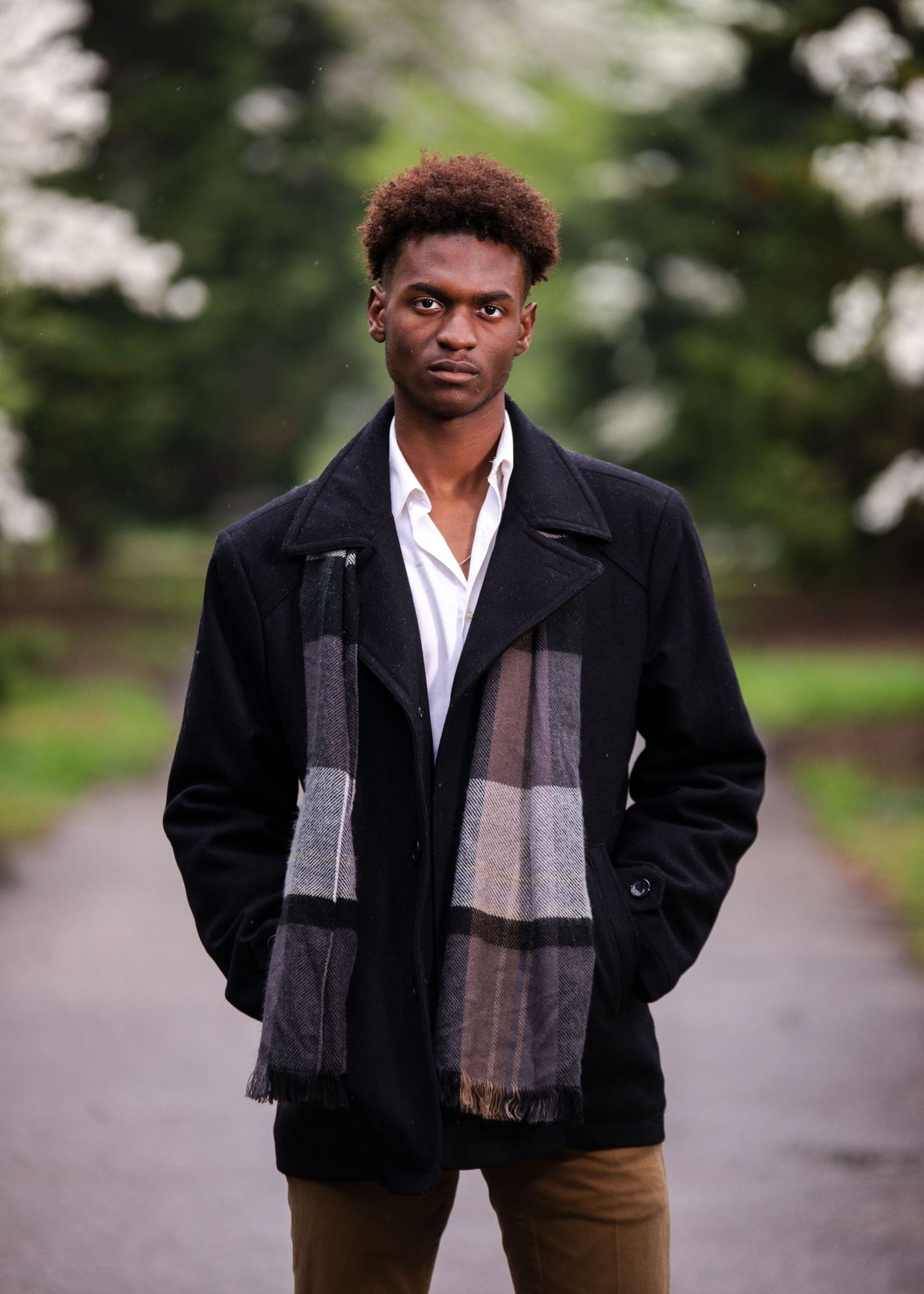 Stylish young Black man in black peacoat and plaid scarf standing on a tree-lined park path.