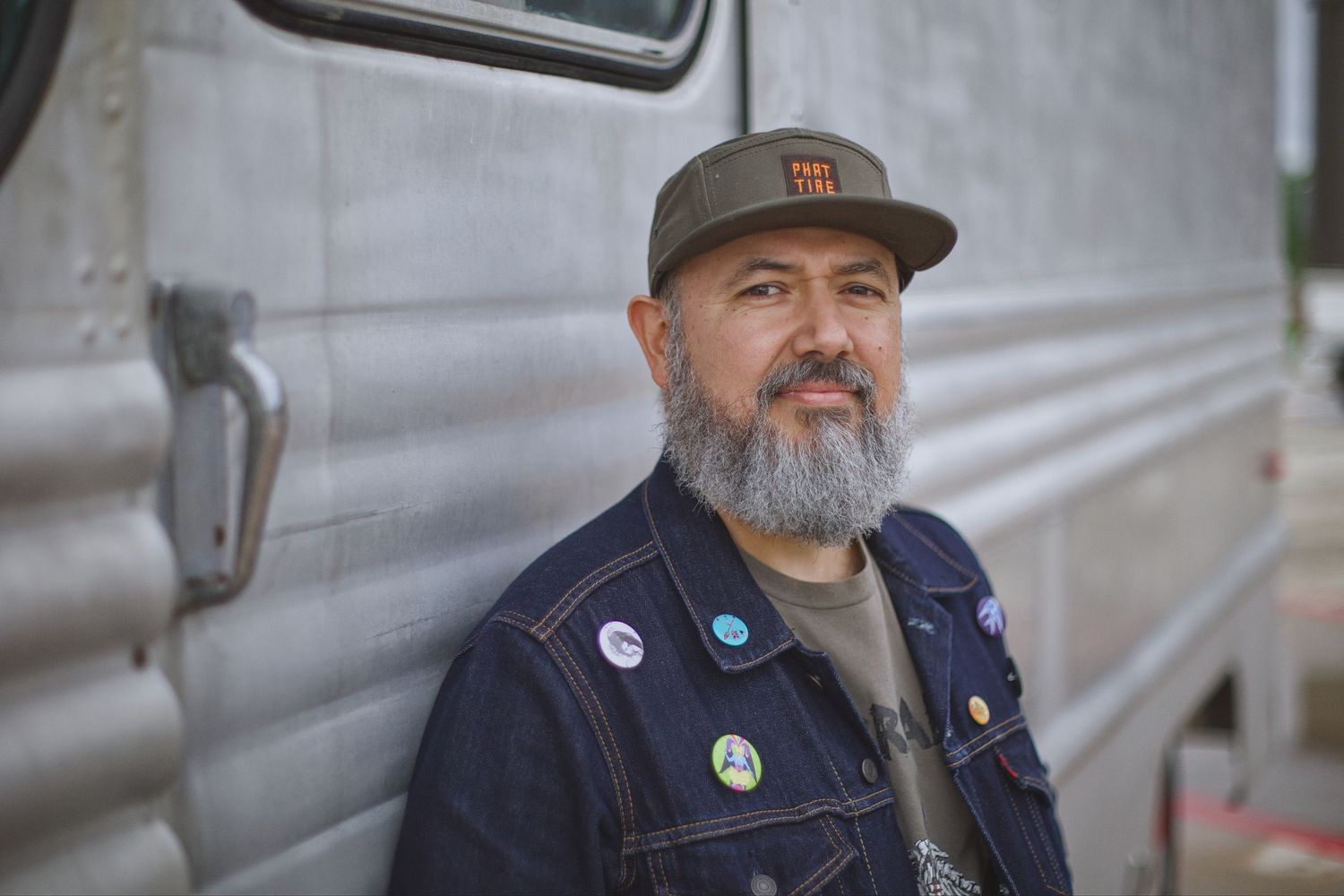 Bearded man in denim jacket with pins and cap poses beside vintage silver trailer outdoors.