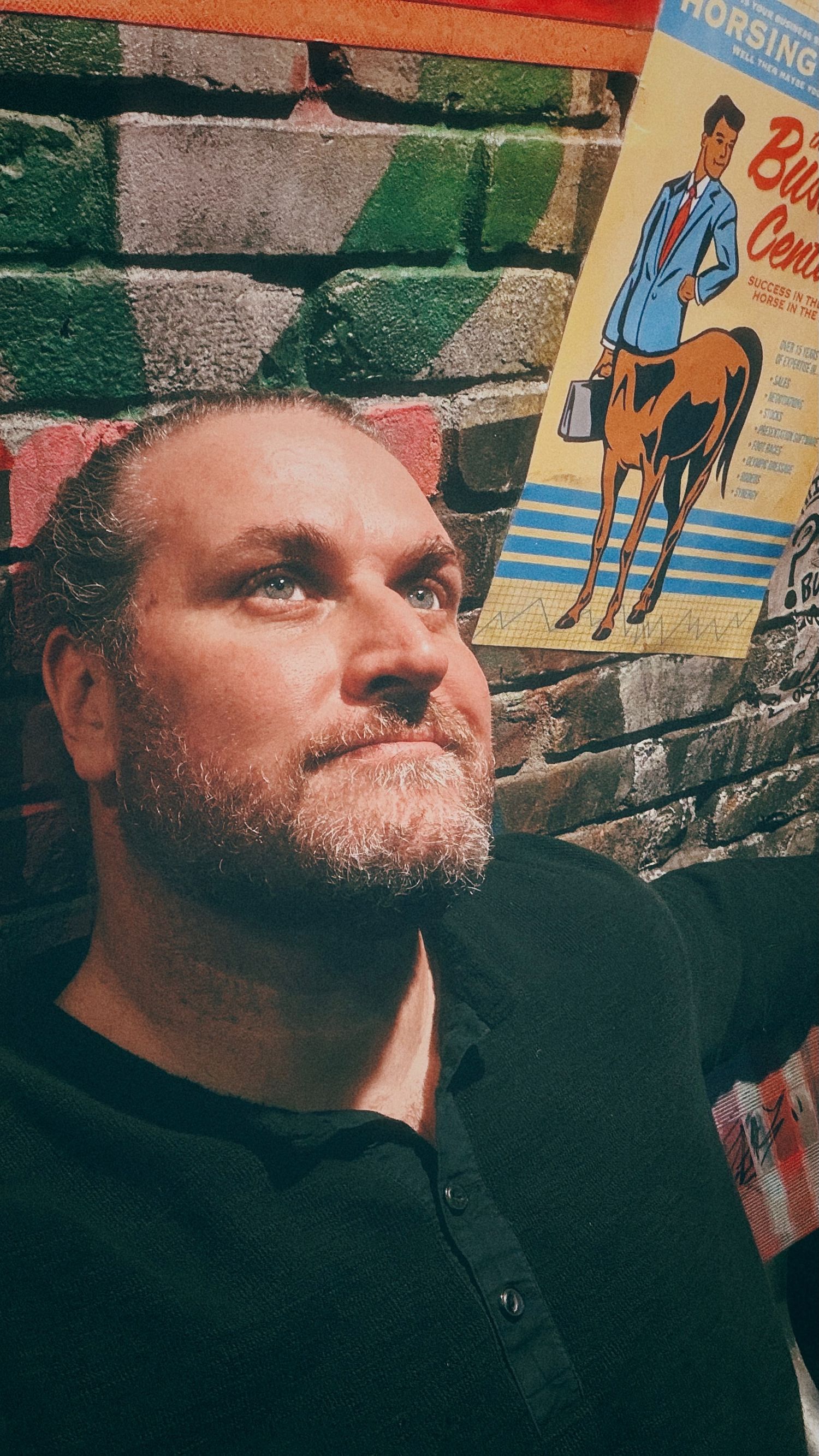 Man with beard gazes upward near vintage cowboy poster on brick wall.