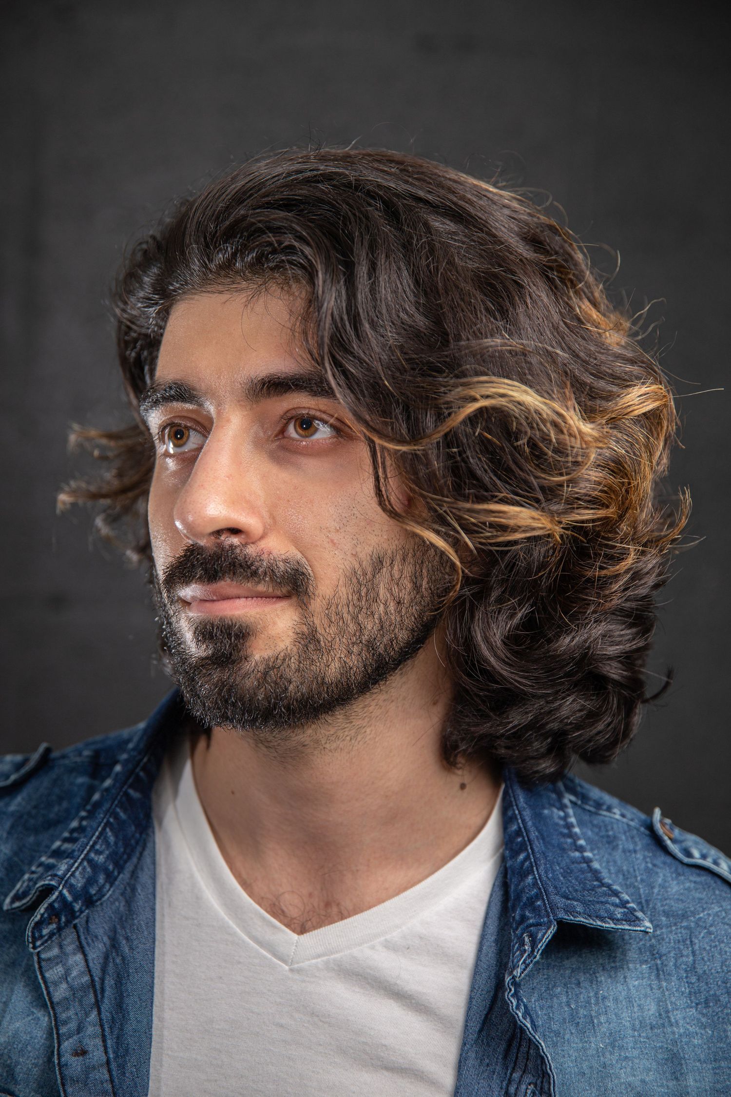 Young man with long wavy dark hair and beard wearing denim jacket and white t-shirt against dark background.
