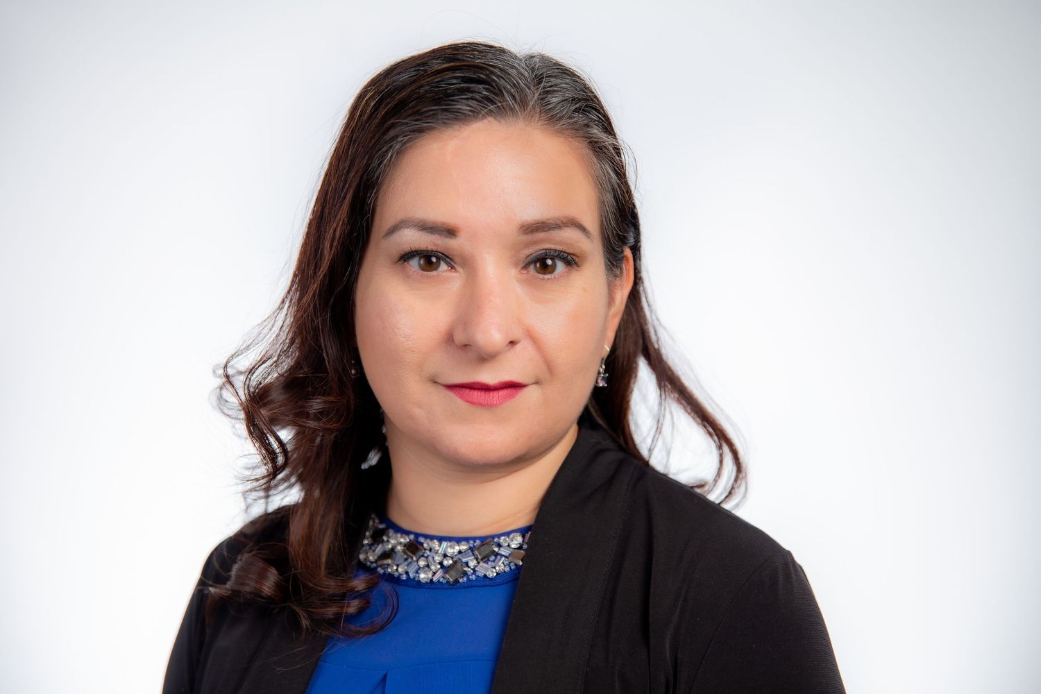 Professional headshot of woman with dark hair wearing blue top and black blazer on white background.