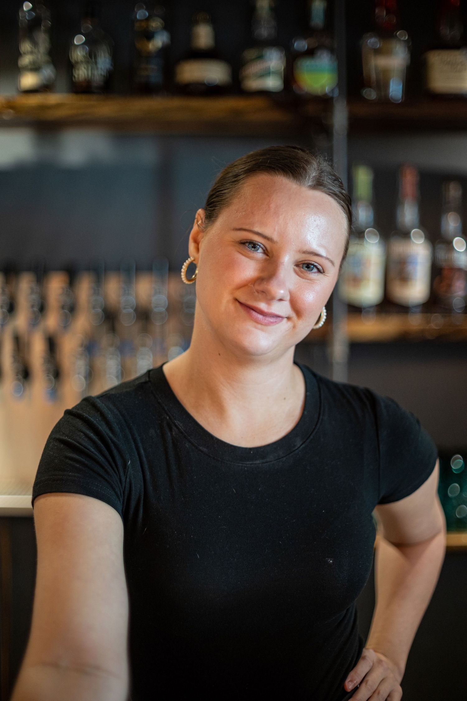 Smiling female bartender in black t-shirt stands confidently in front of a well-stocked bar.