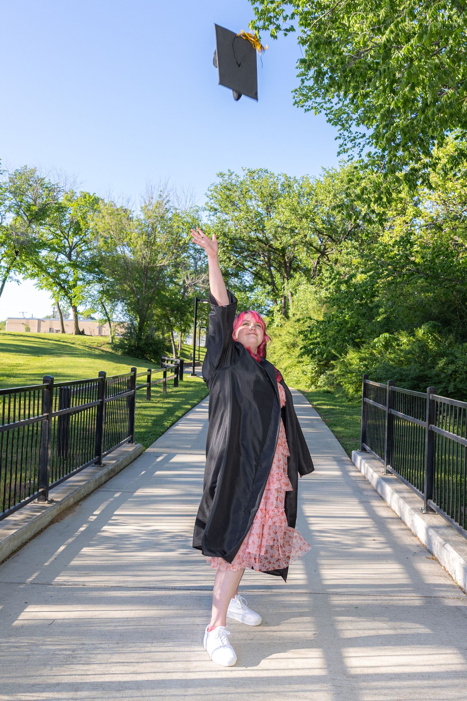 Young woman in black graduation gown throws cap in air on bridge surrounded by lush green trees.