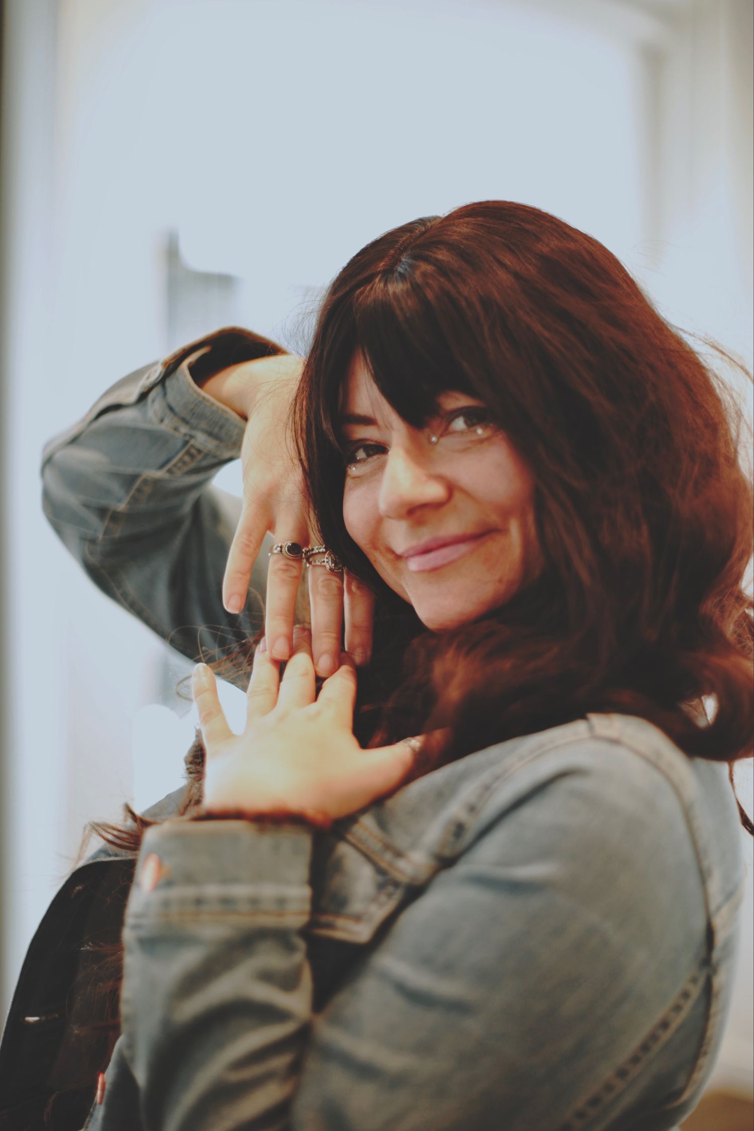 Smiling brunette woman in denim jacket with rings on her fingers posing indoors.