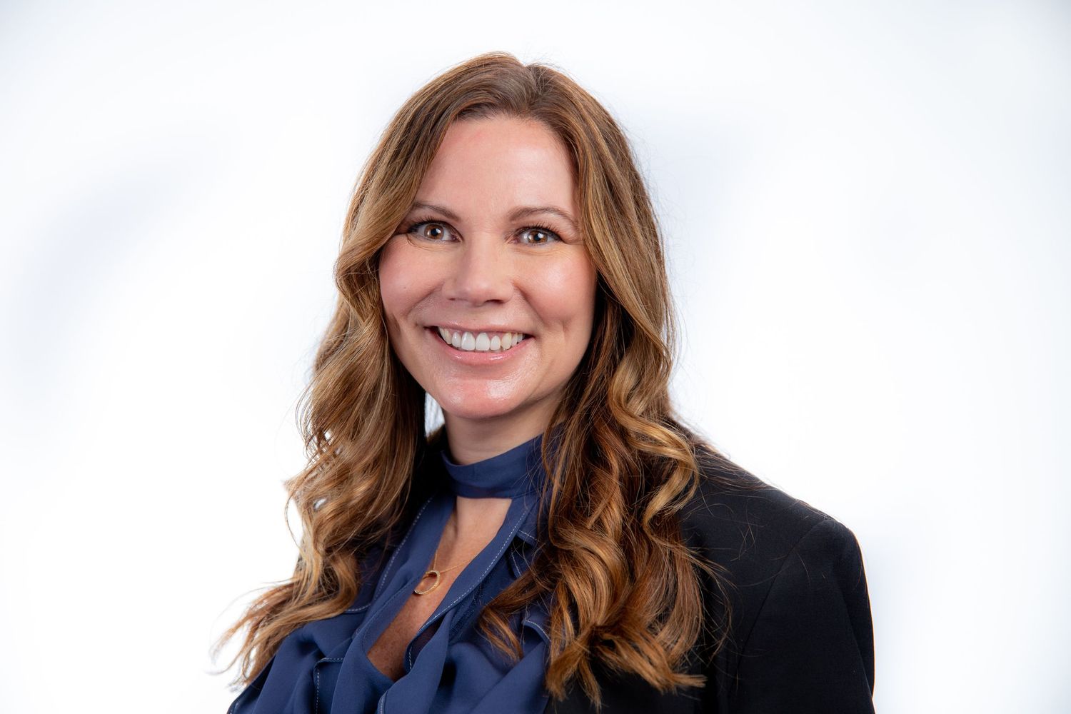 Smiling woman with wavy brown hair wearing a blue blouse and black blazer on white background.