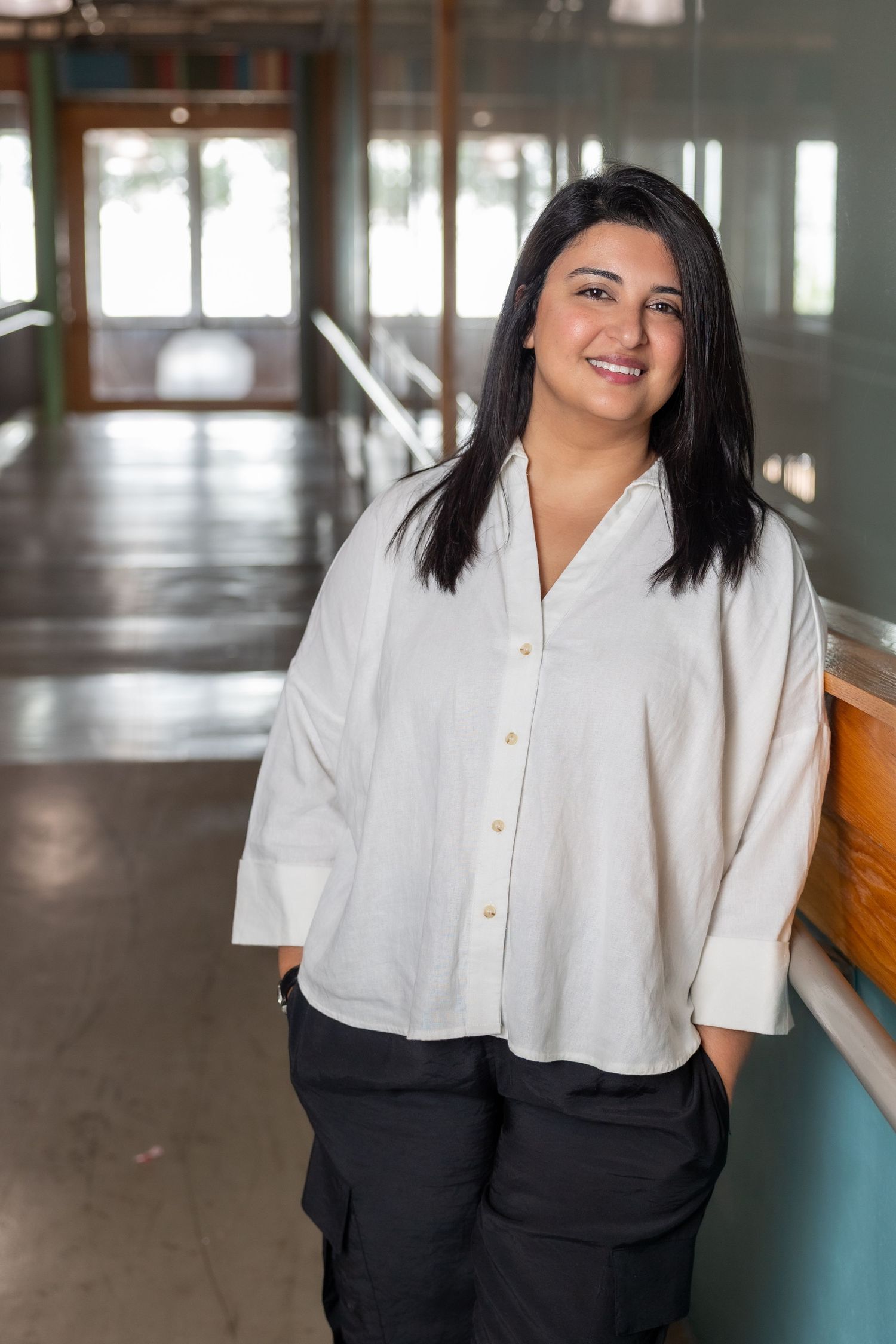 Woman with black hair smiling in a white button-up shirt and black pants in a modern hallway.