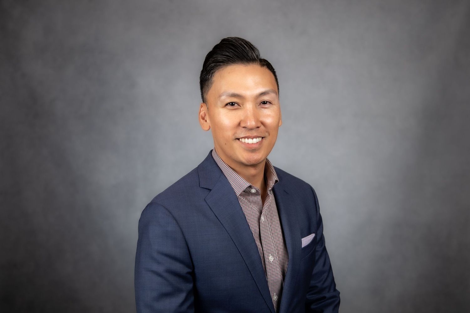 Professional headshot of a smiling Asian man in a navy blue blazer and gray shirt against a gray background.