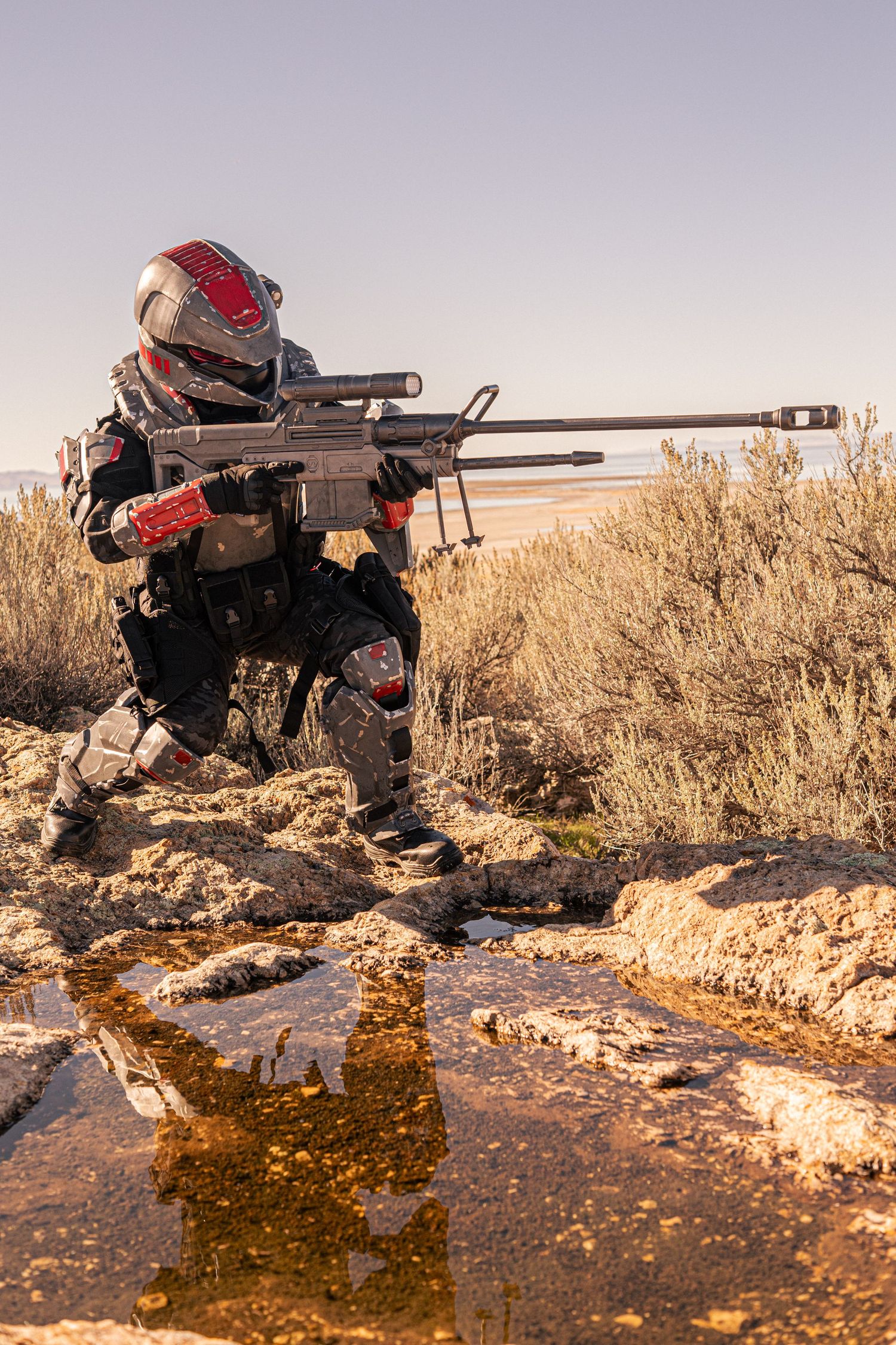 Cosplayer in futuristic armor aims a sniper rifle while crouching near a muddy stream in a desert landscape.