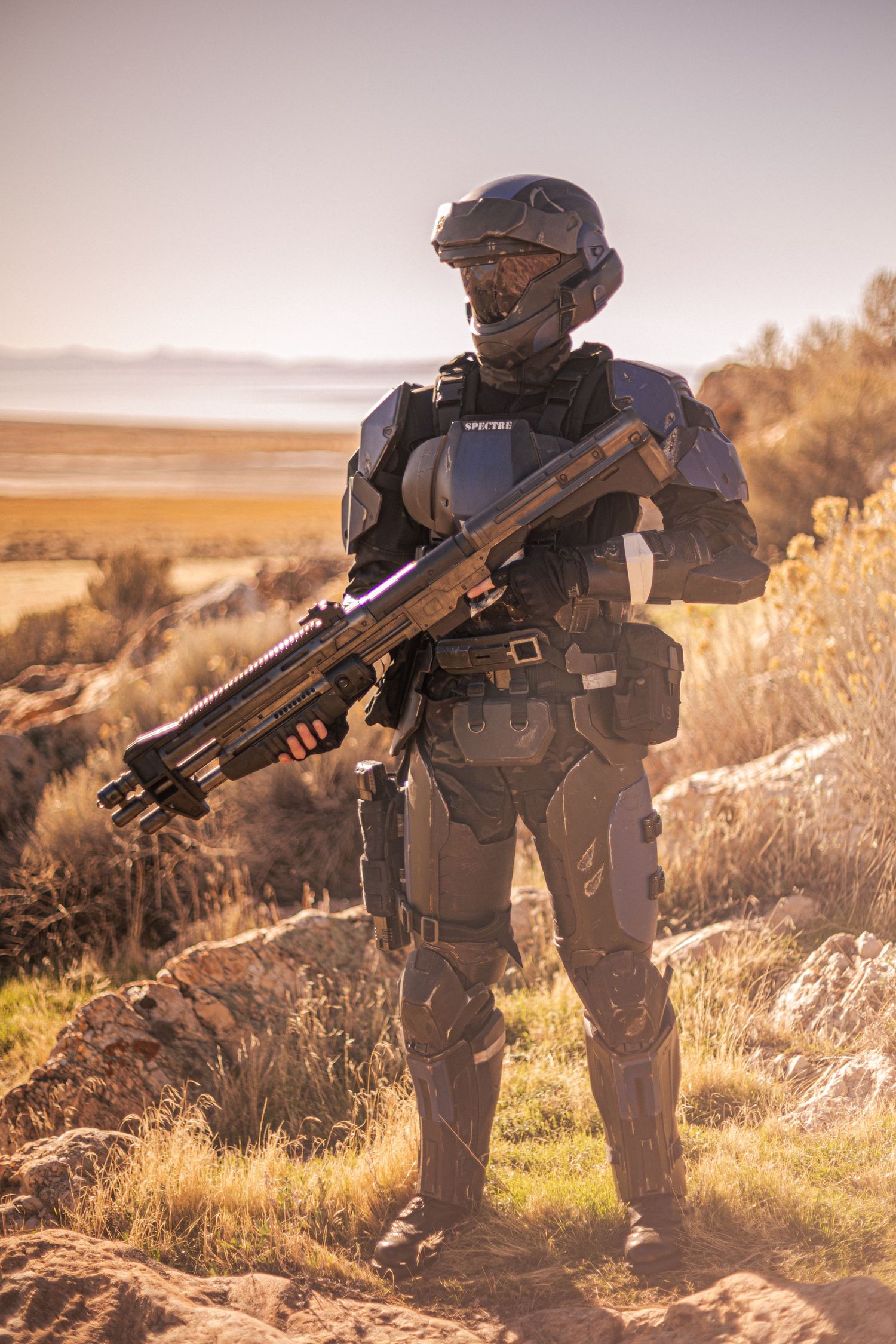 Halo ODST cosplayer in SPECTRE armor holds large prop weapon on rocky desert terrain.