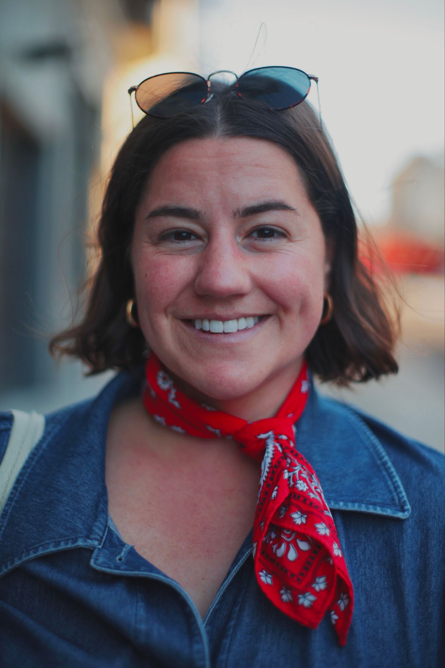 Smiling woman with sunglasses on head wearing red bandana and denim jacket outdoors.
