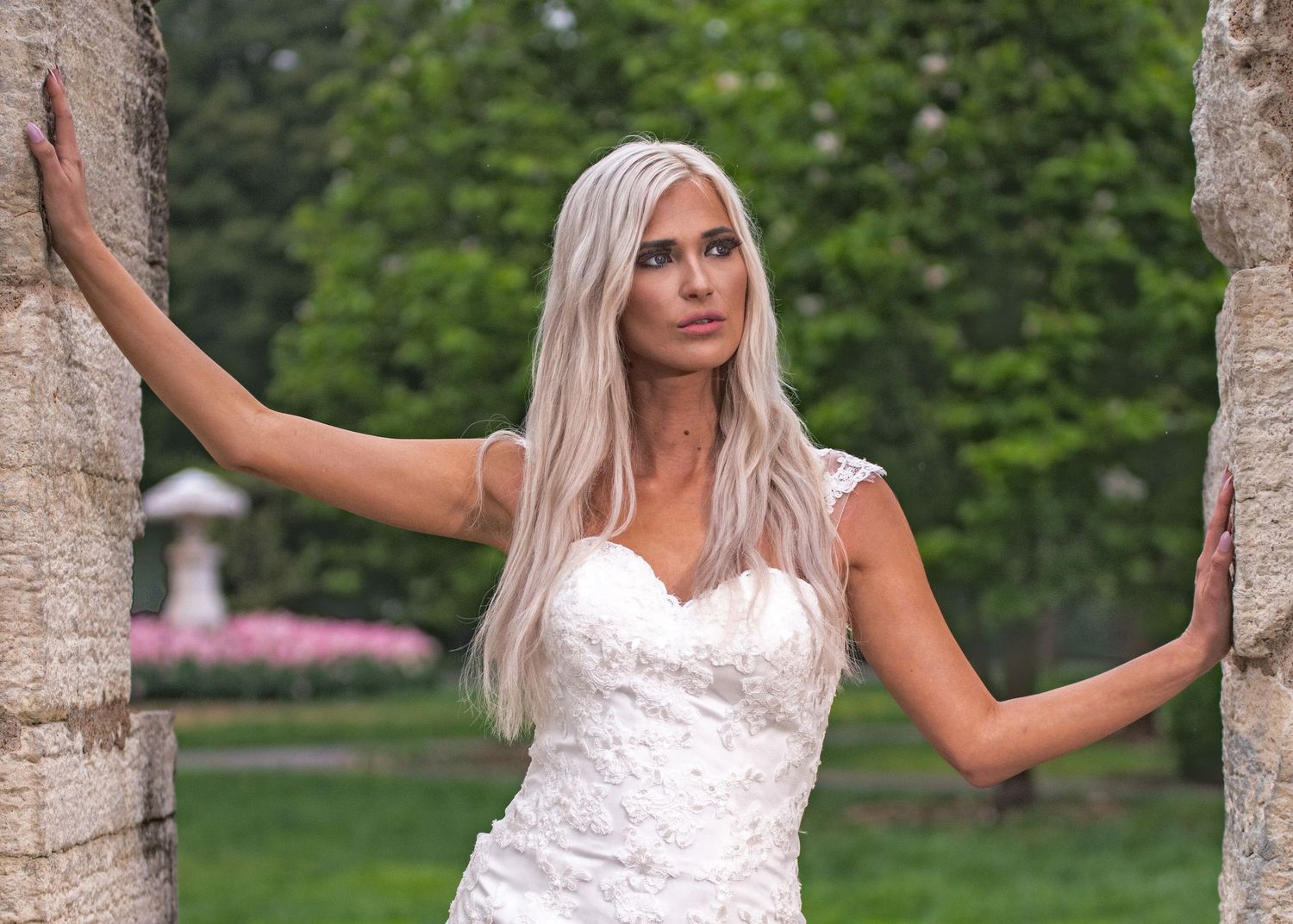 Blonde woman in white lace wedding dress posing between two trees in a lush green garden.