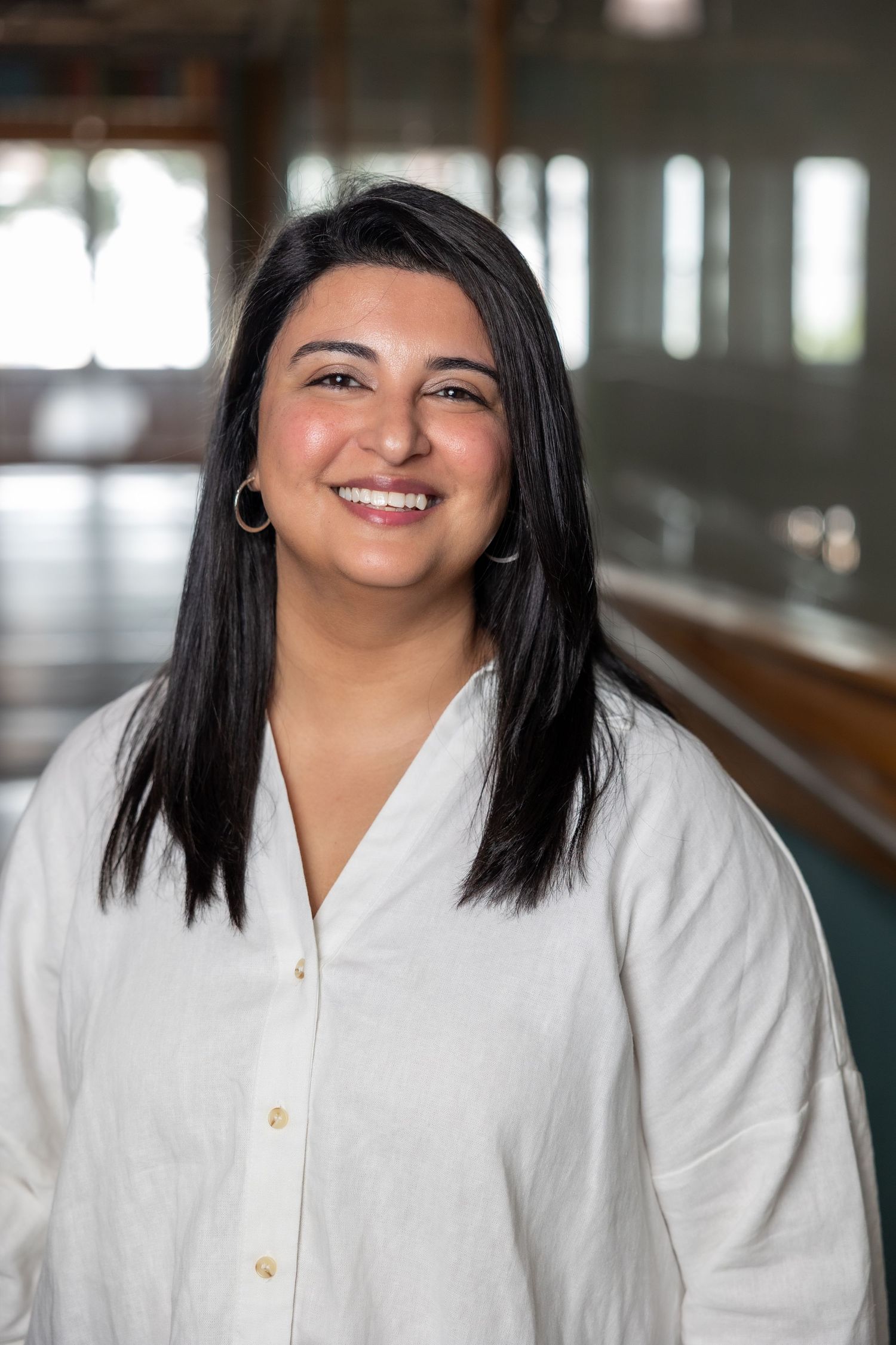 Smiling woman with black hair wearing a white linen shirt, gold hoop earrings, in a bright indoor setting.