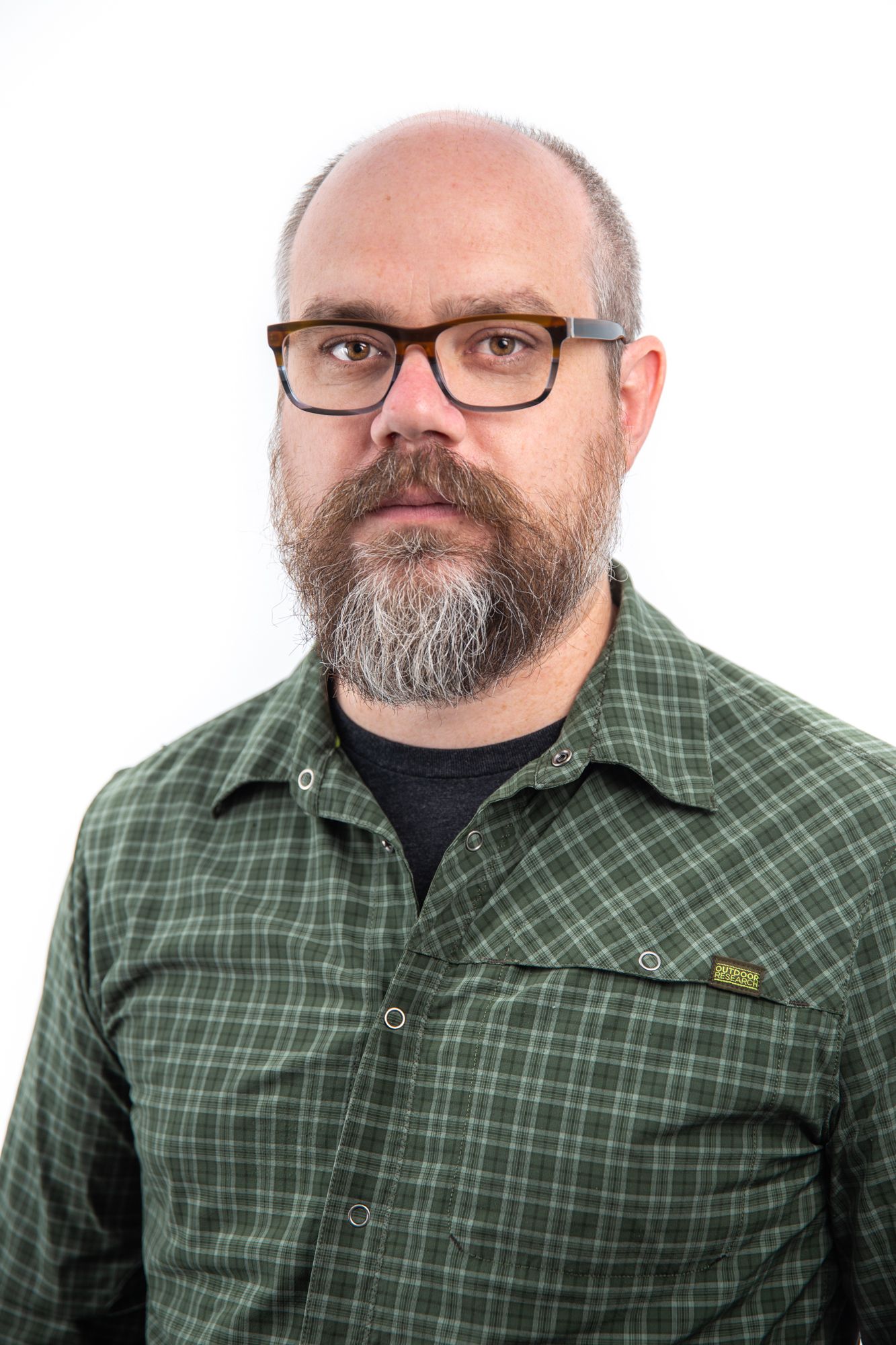 Bald man with gray beard wearing glasses and green plaid shirt against white background.