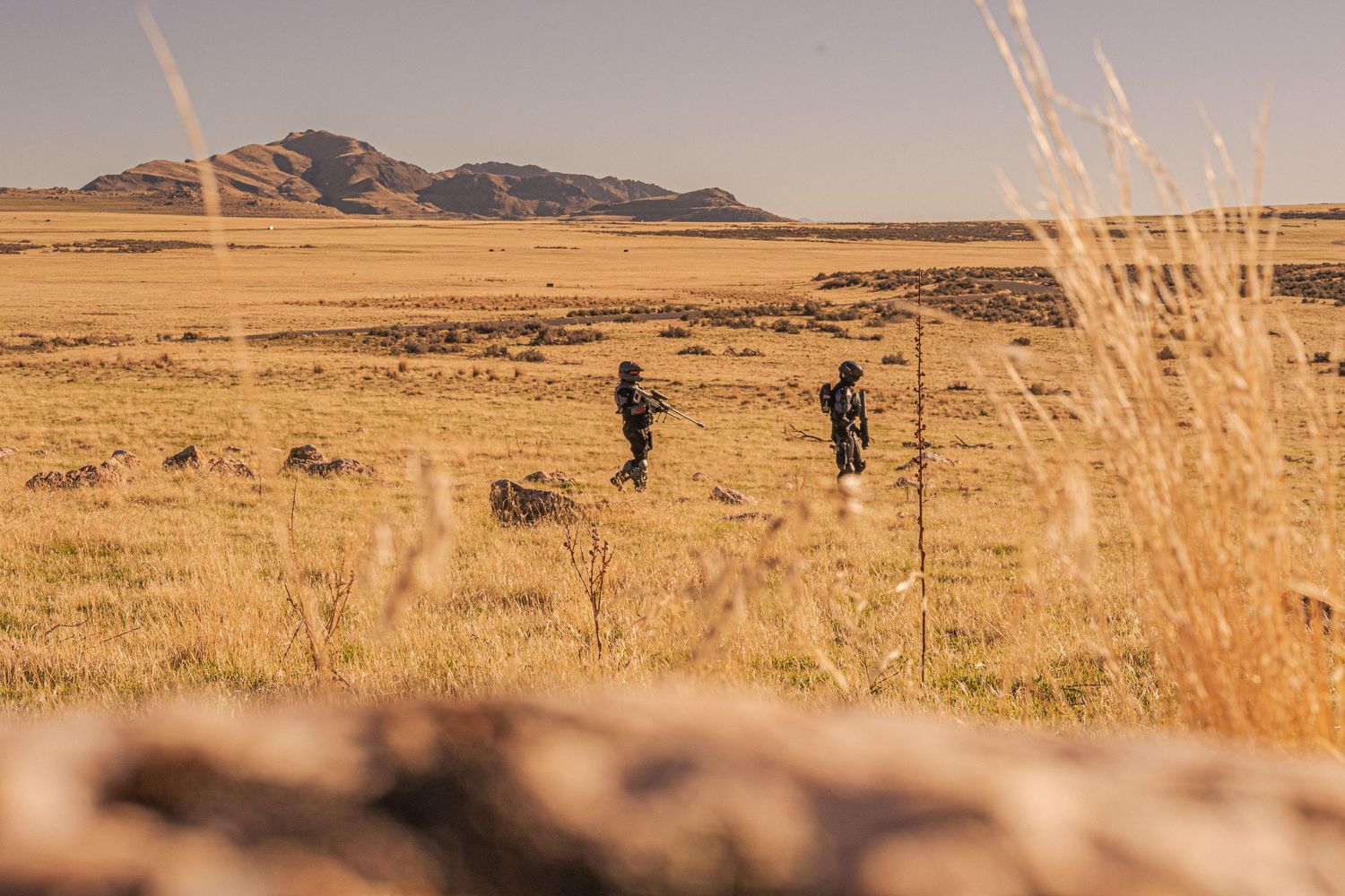 Two soldiers in tactical gear standing in a vast arid desert landscape with mountains behind.