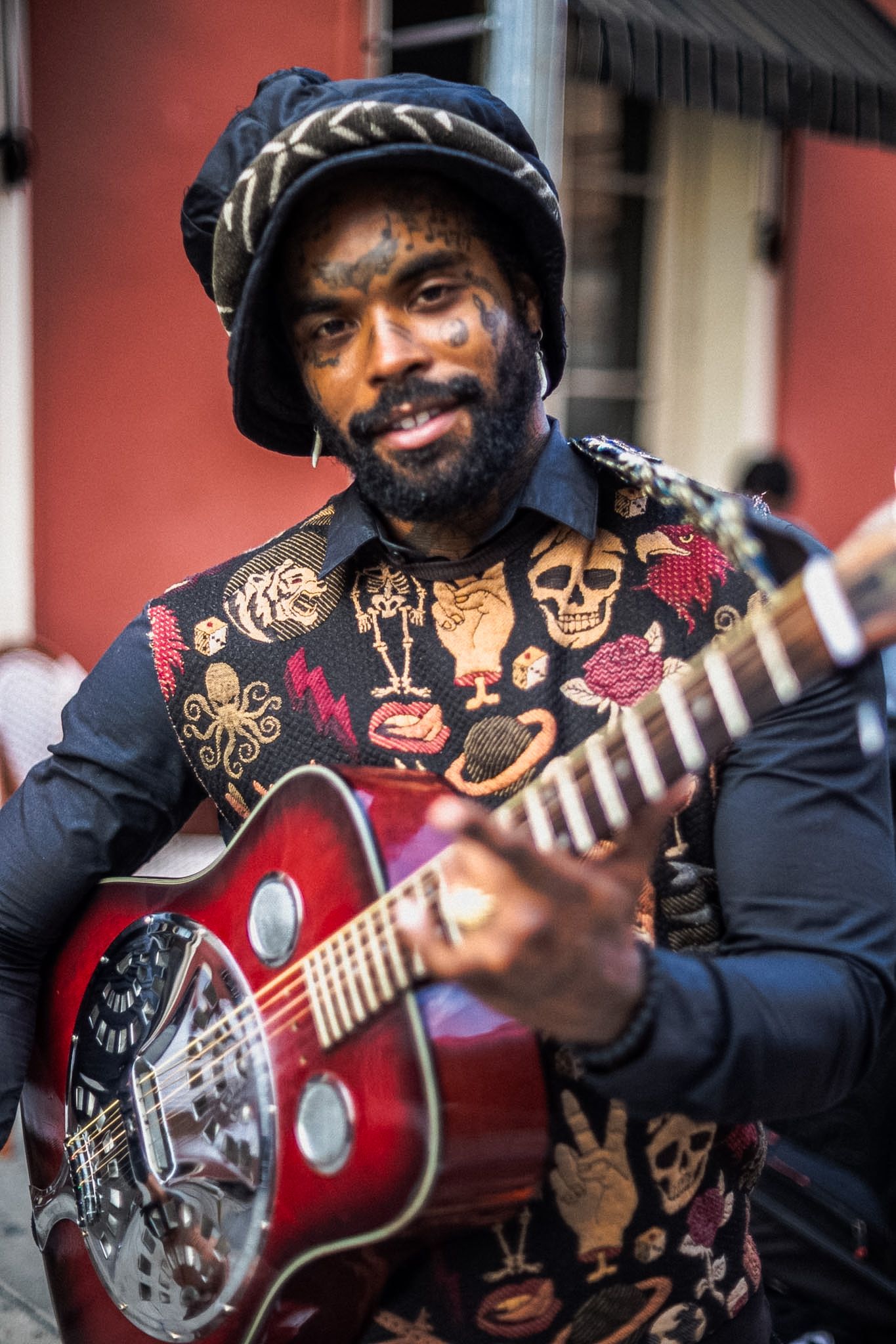 Smiling street musician playing a red resonator guitar, wearing a decorative vest with skull patches and a black hat.