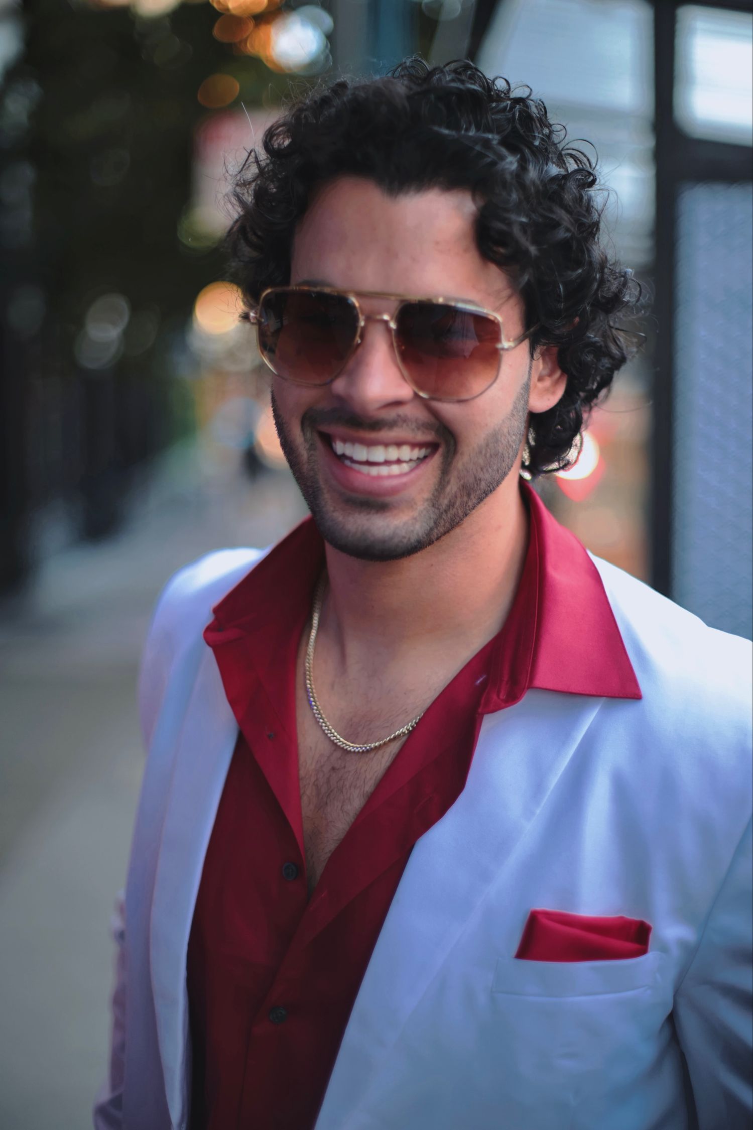 Handsome man with curly hair in white blazer and red shirt smiling on city sidewalk.
