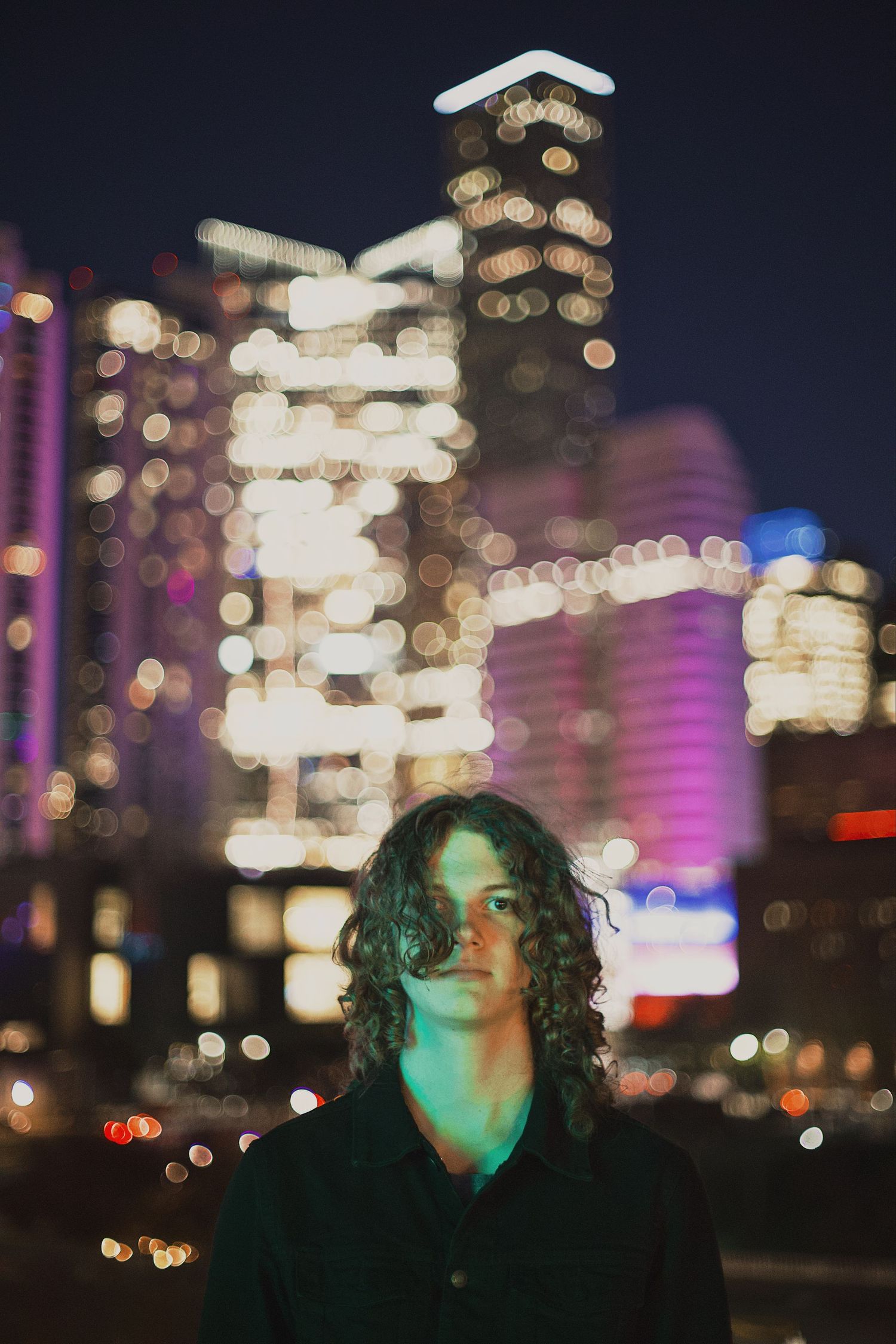 Young man with curly hair standing on a rooftop at night with city lights bokeh background.