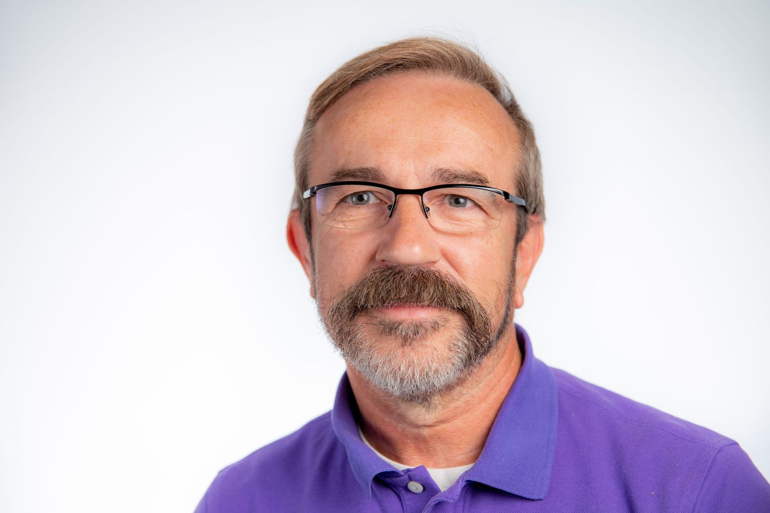 Professional headshot of bearded man wearing glasses and purple polo shirt on white background.
