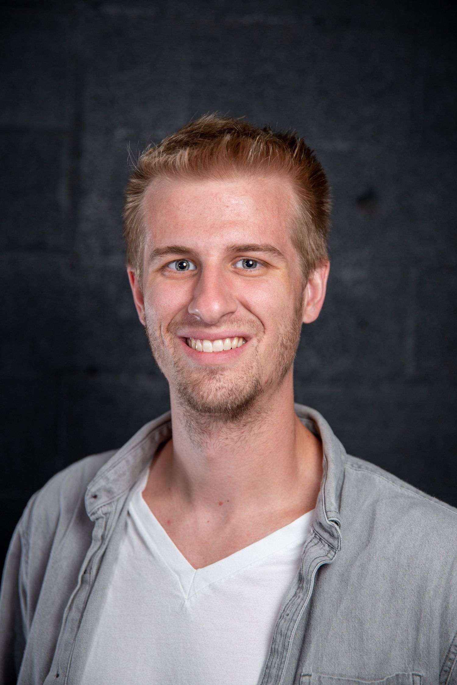 Smiling young man with blonde hair wearing a gray shirt over white t-shirt against dark background.