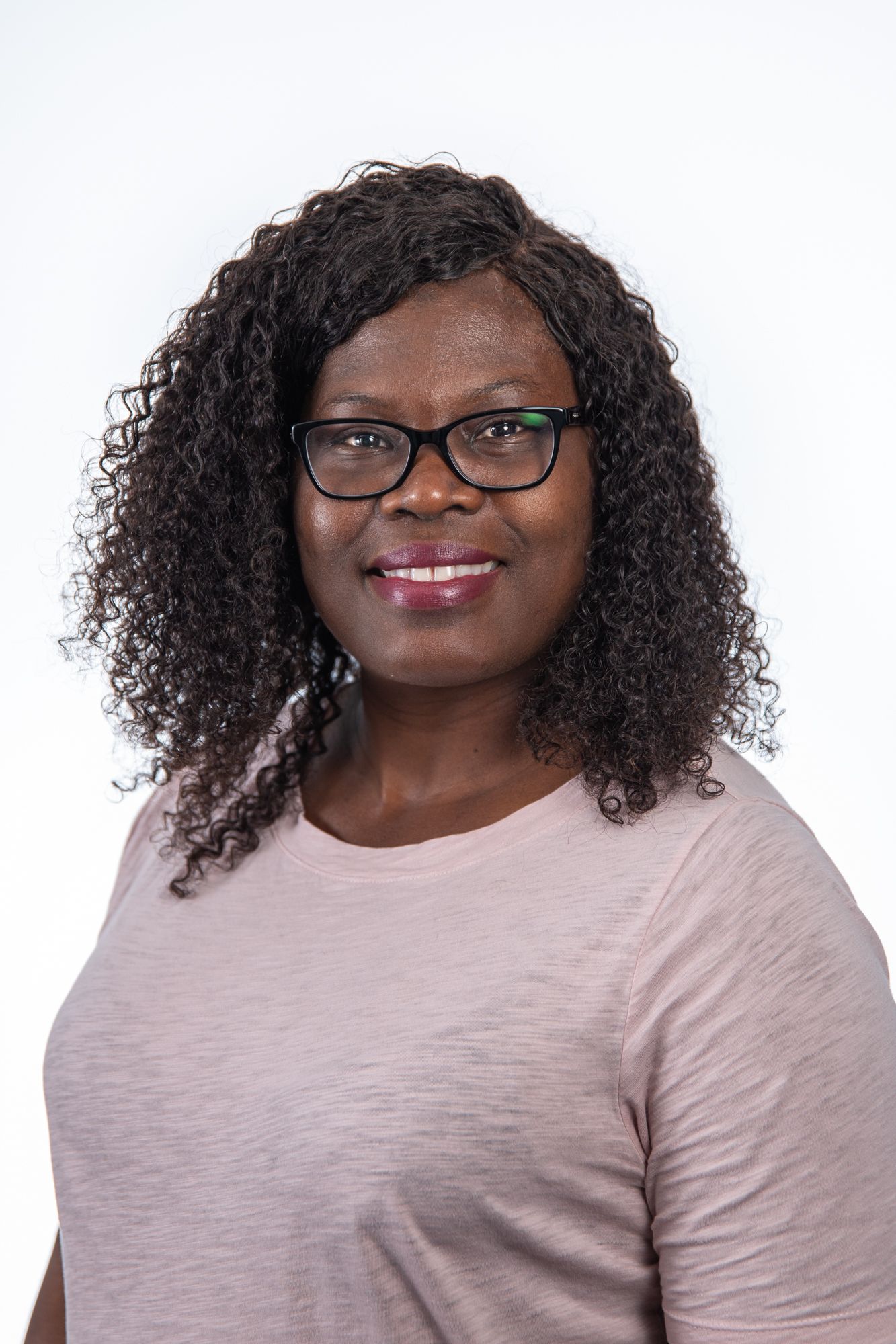 Smiling woman with curly black hair wearing glasses and pink top on white background.