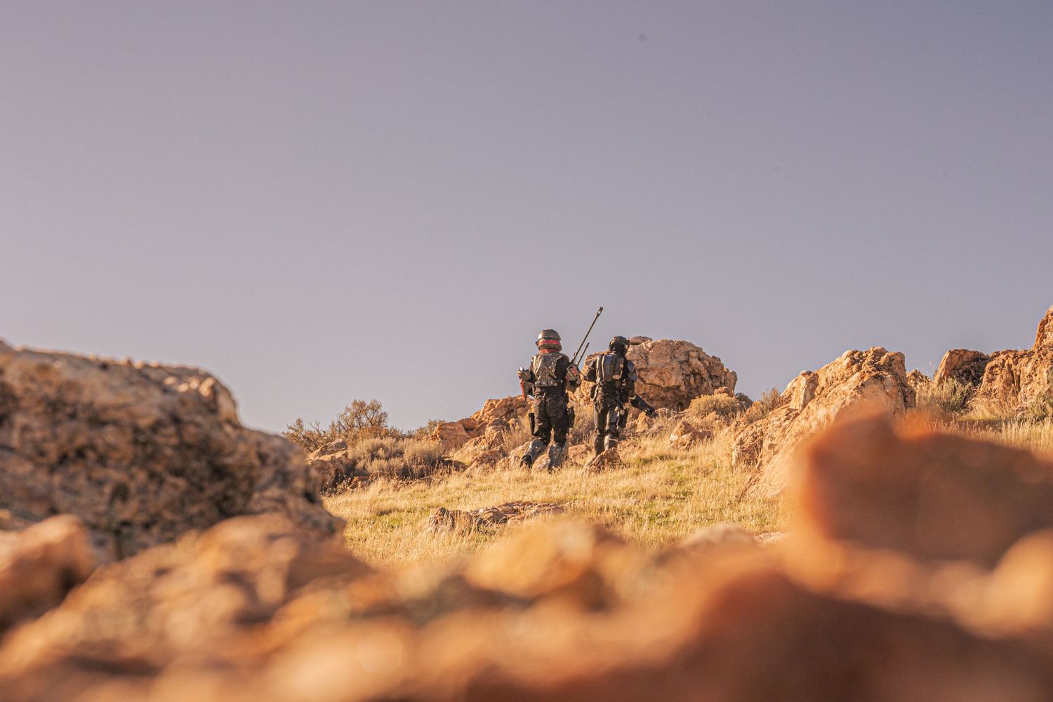 Two armed figures in black tactical gear silhouetted against rocky desert terrain under a clear sky.