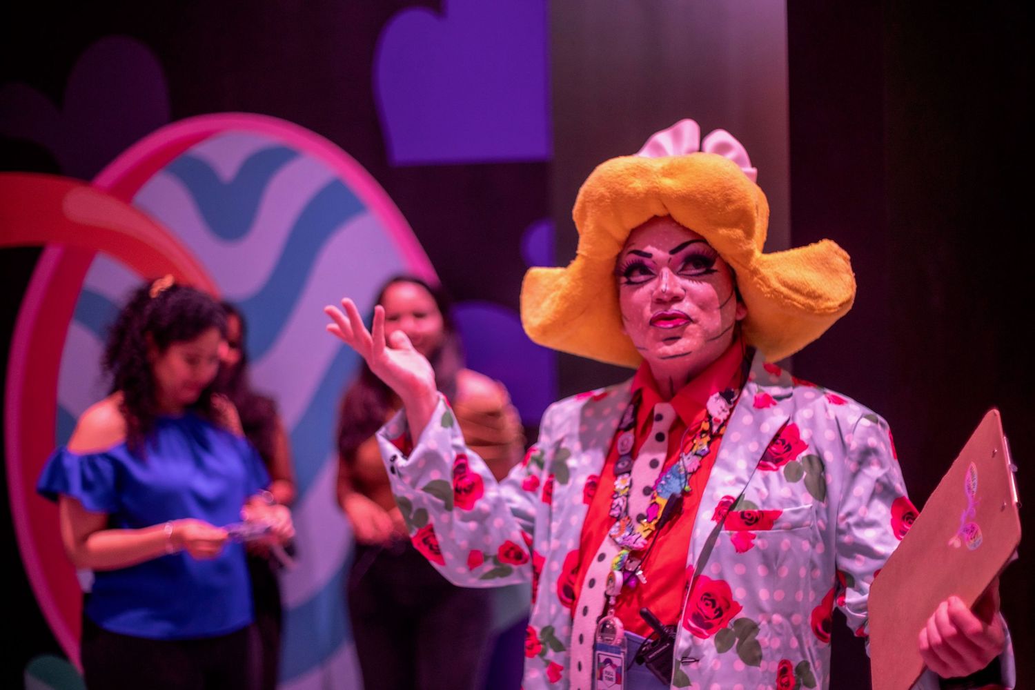 Performer in colorful floral costume and yellow hat gestures dramatically on stage during theater production.