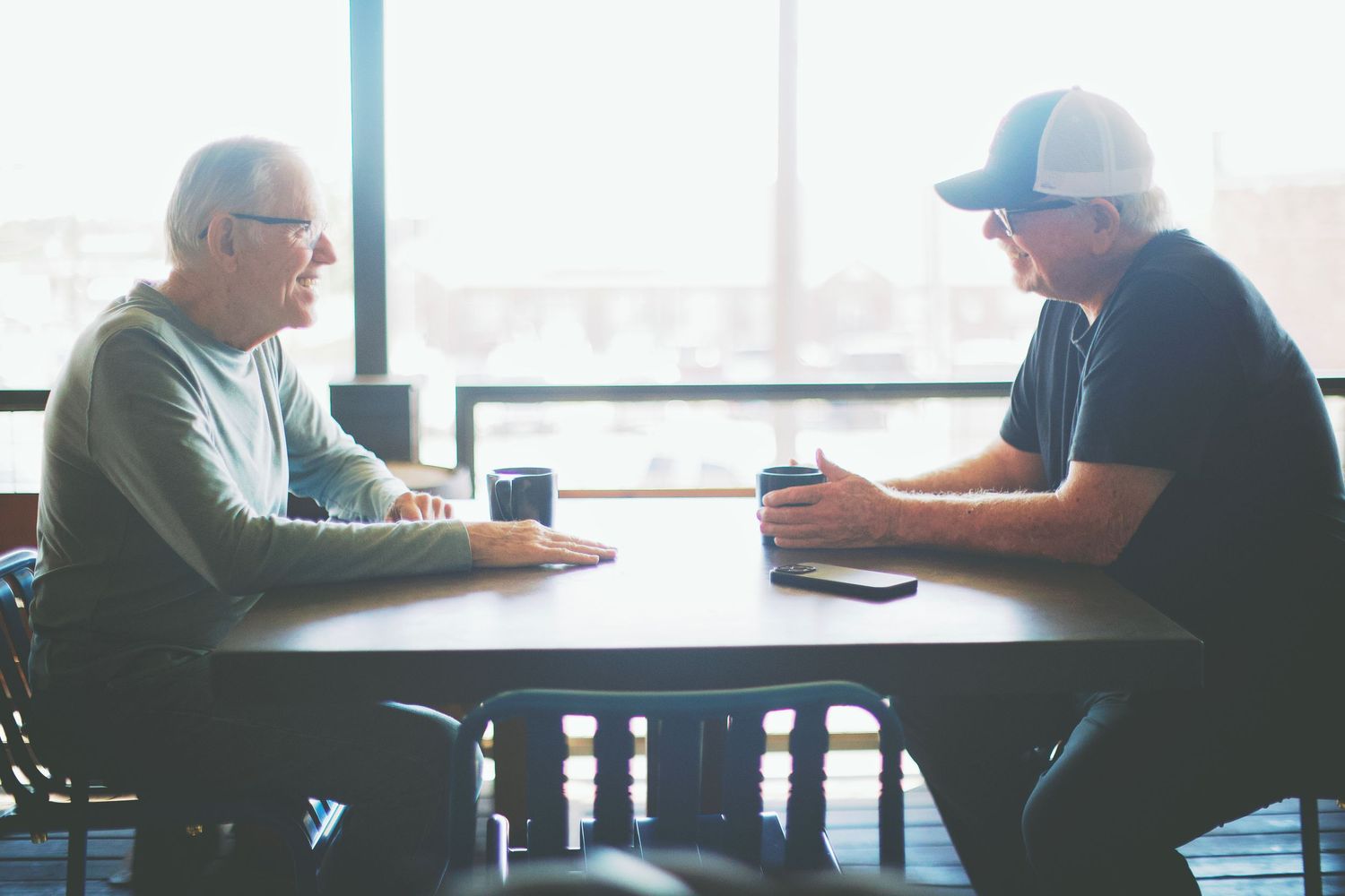 Two men sitting across from each other at a wooden table having a conversation near a bright window.