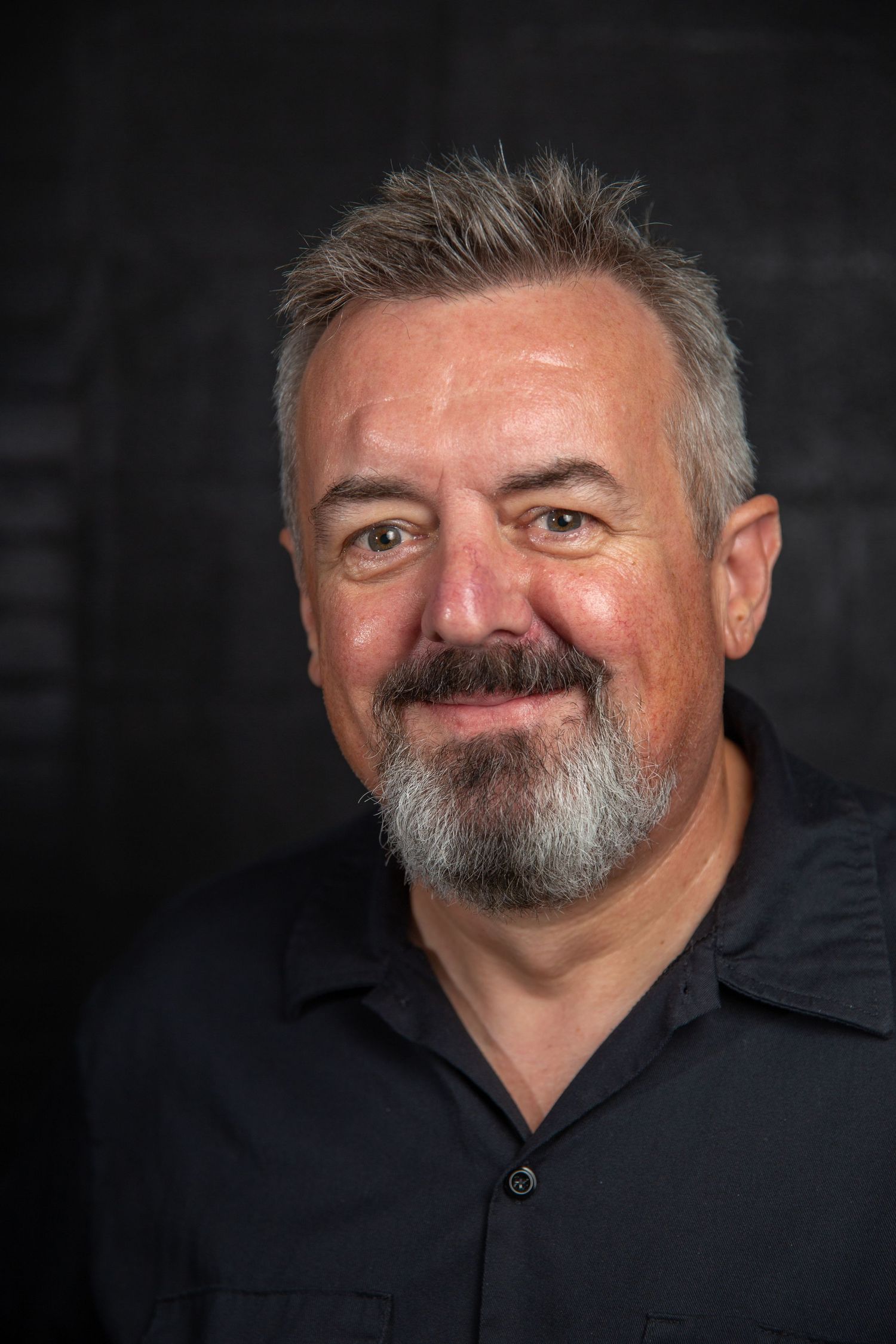 Middle-aged man with gray beard smiling warmly wearing black shirt against dark background.