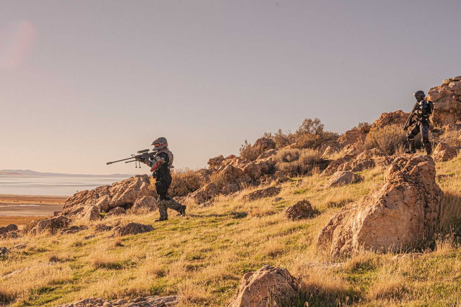 Two armed soldiers in tactical gear survey a rocky hillside overlooking a vast open landscape at dusk.