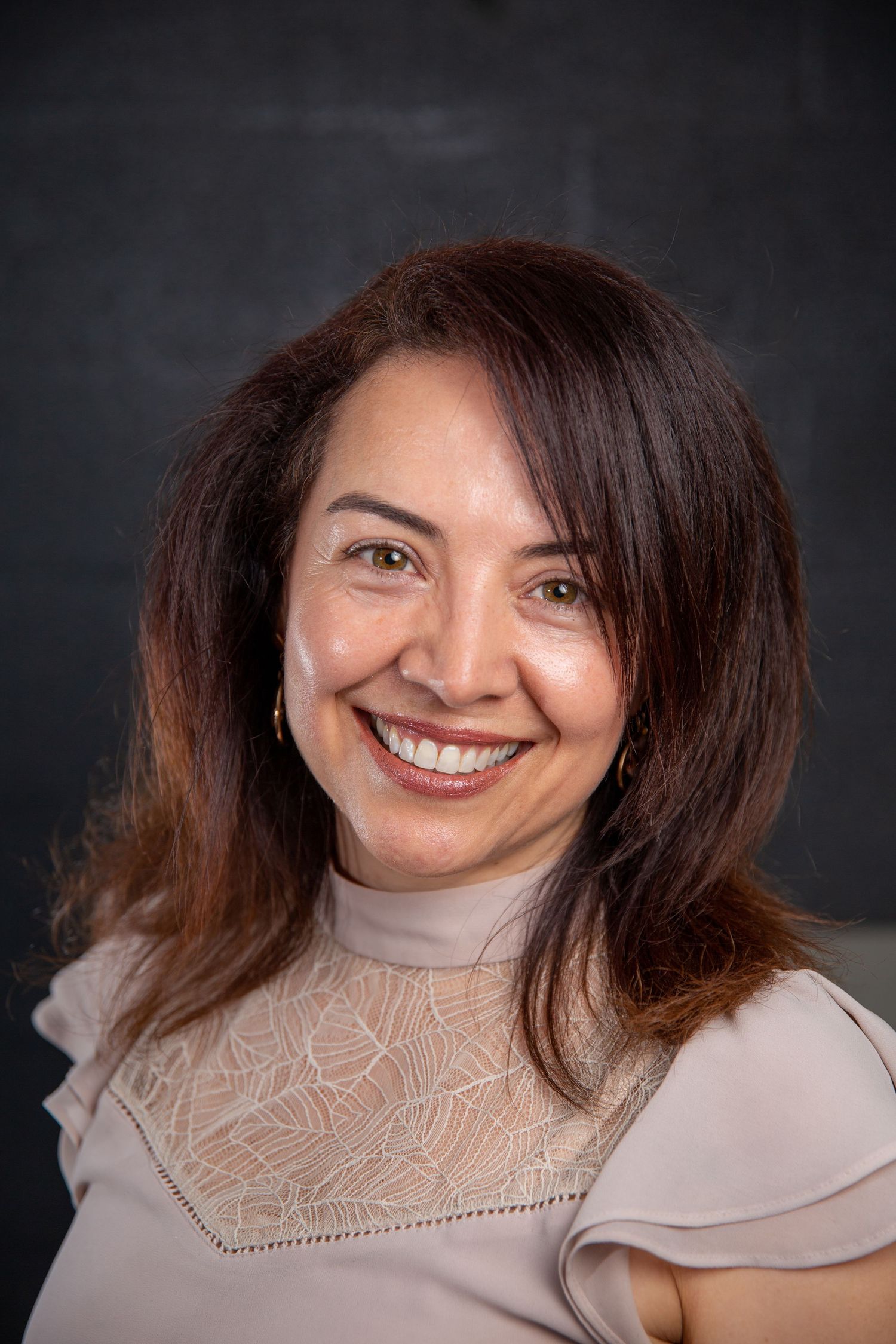Smiling Hispanic woman with brown hair wearing beige lace top against dark background.