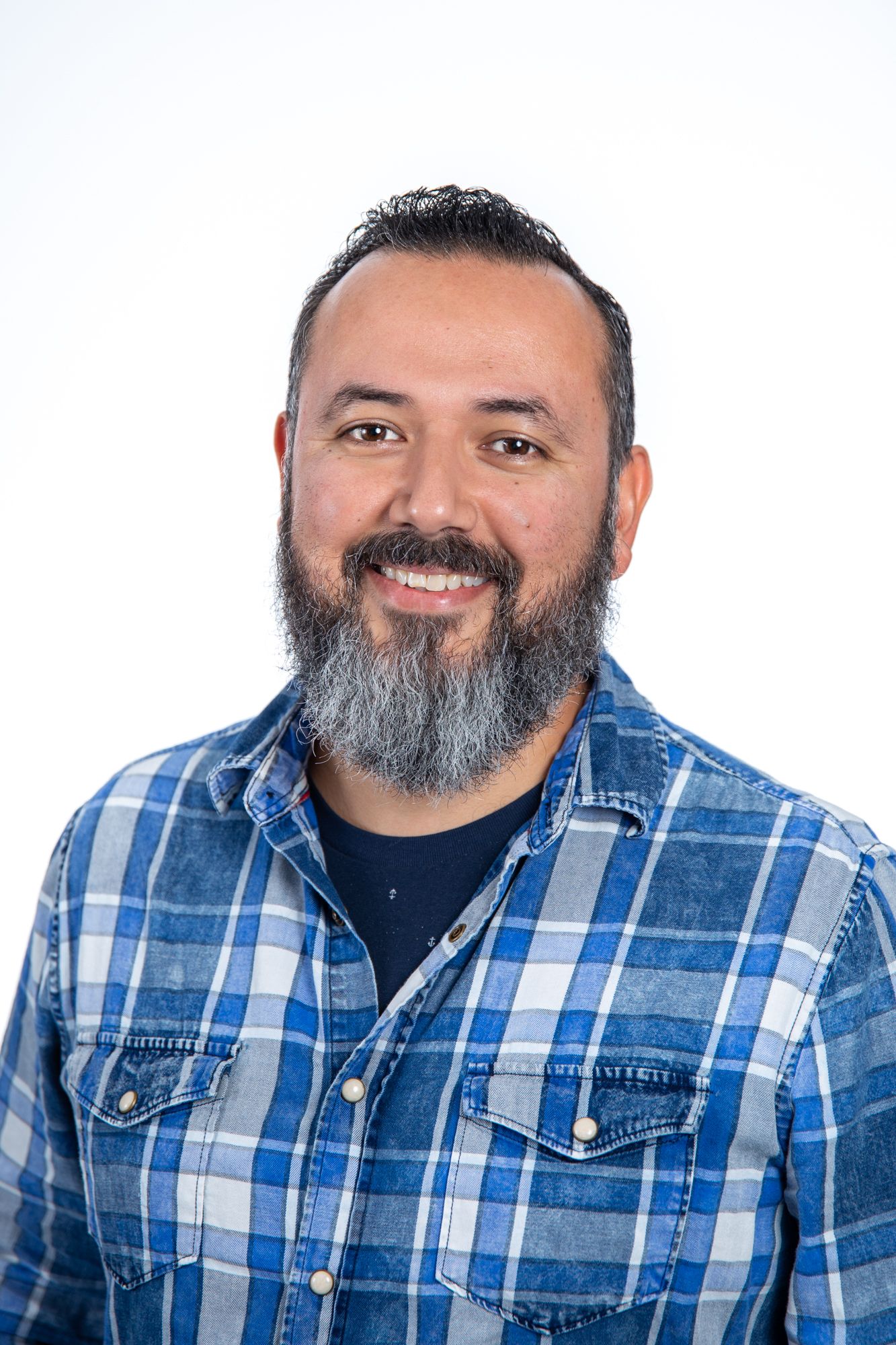 Smiling man with black hair and gray beard wearing a blue plaid shirt, professional headshot on white background.