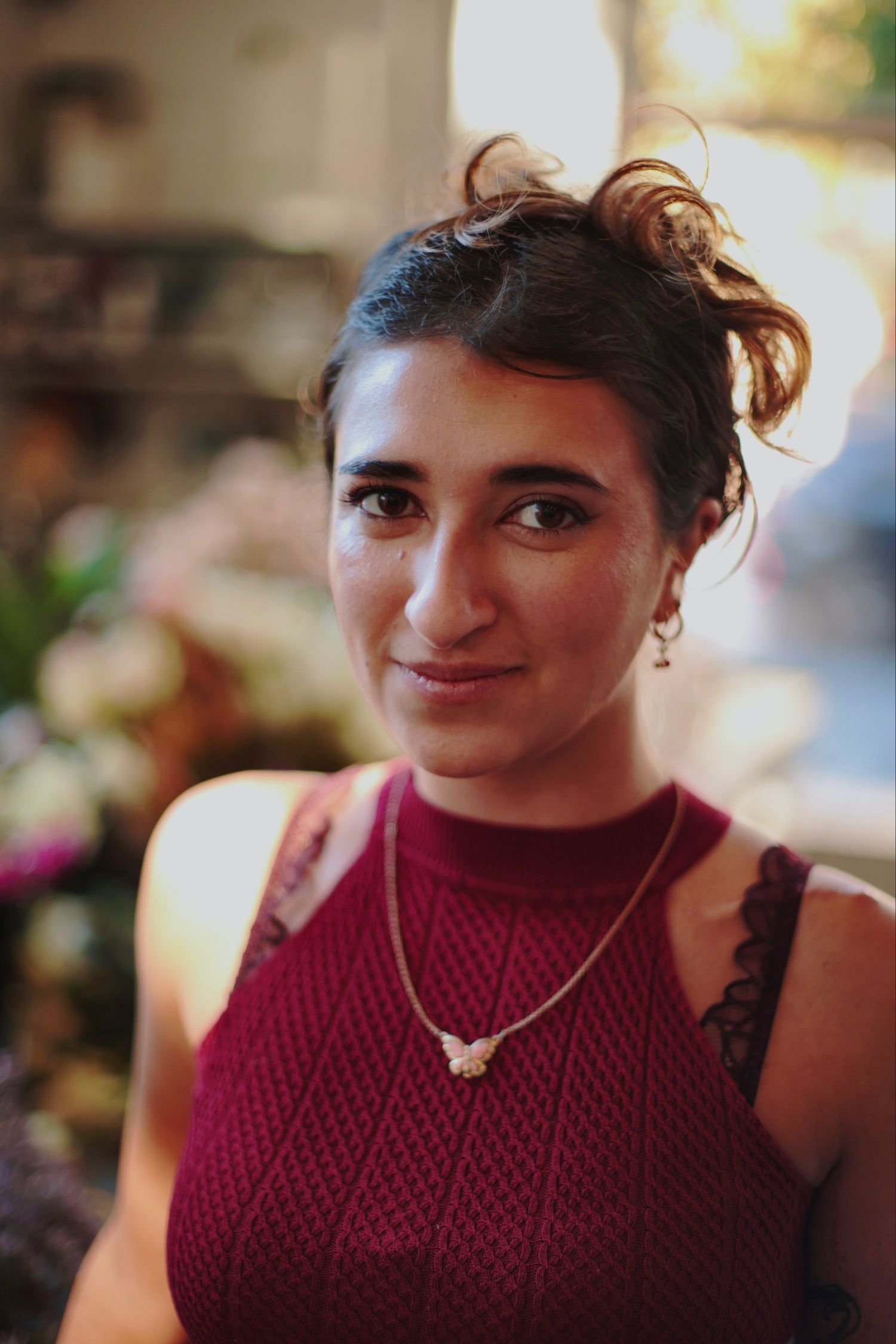 Young woman in burgundy sleeveless top with necklace smiling softly in warm backlit setting.