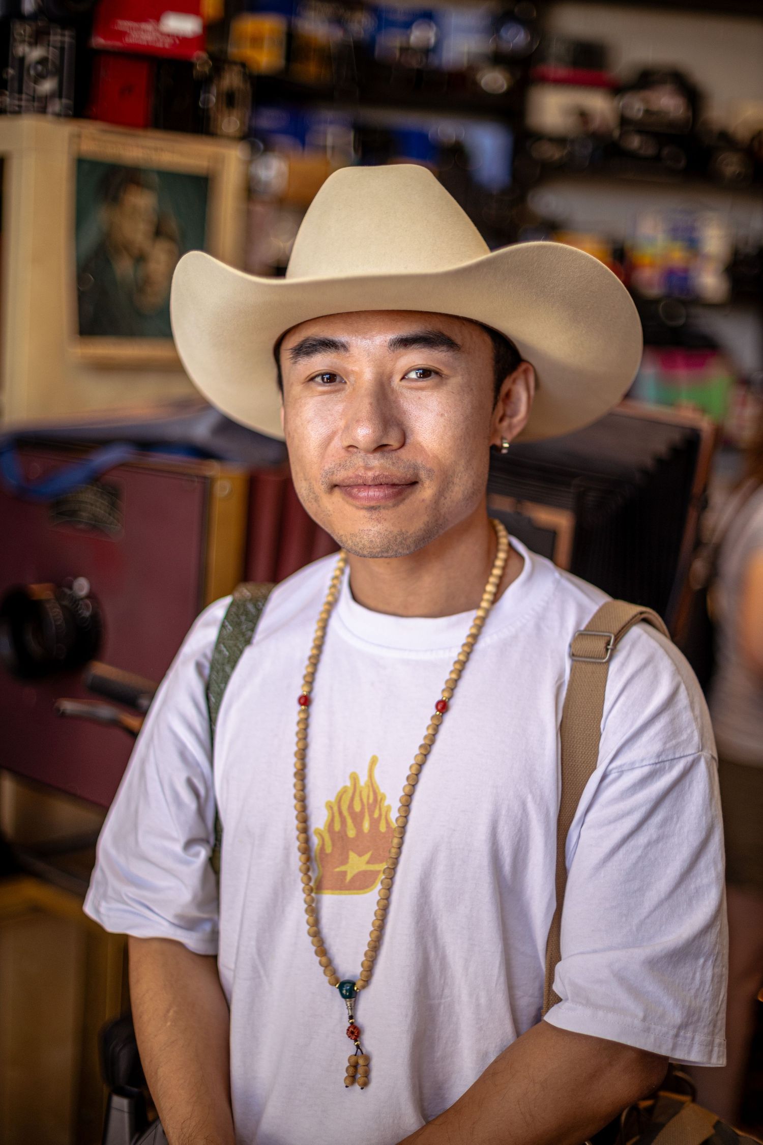 Young Asian man wearing cowboy hat and beaded necklace smiles inside a vintage shop.