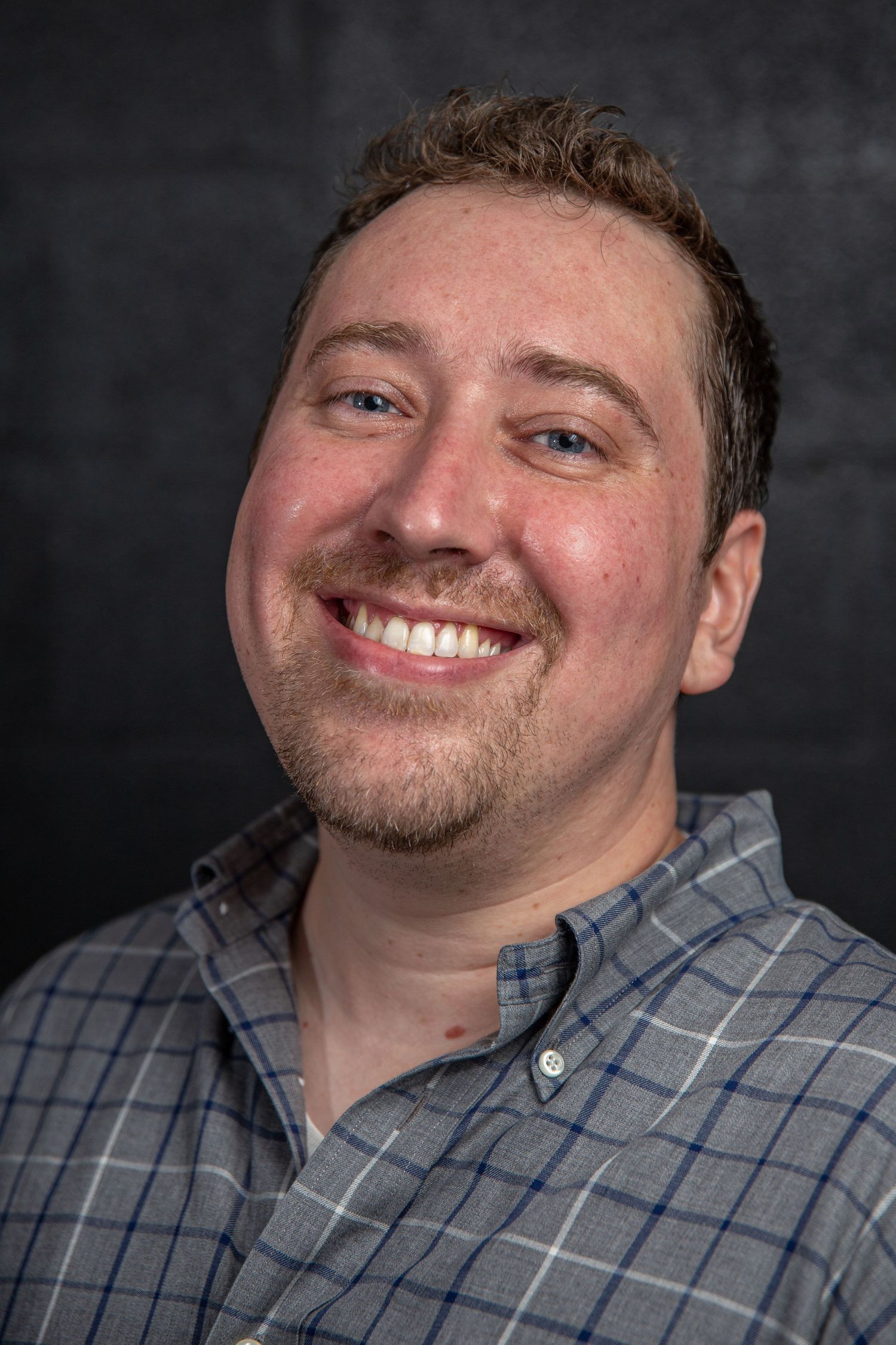 Cheerful man with short brown hair and beard wearing a plaid shirt, professional headshot on dark background.