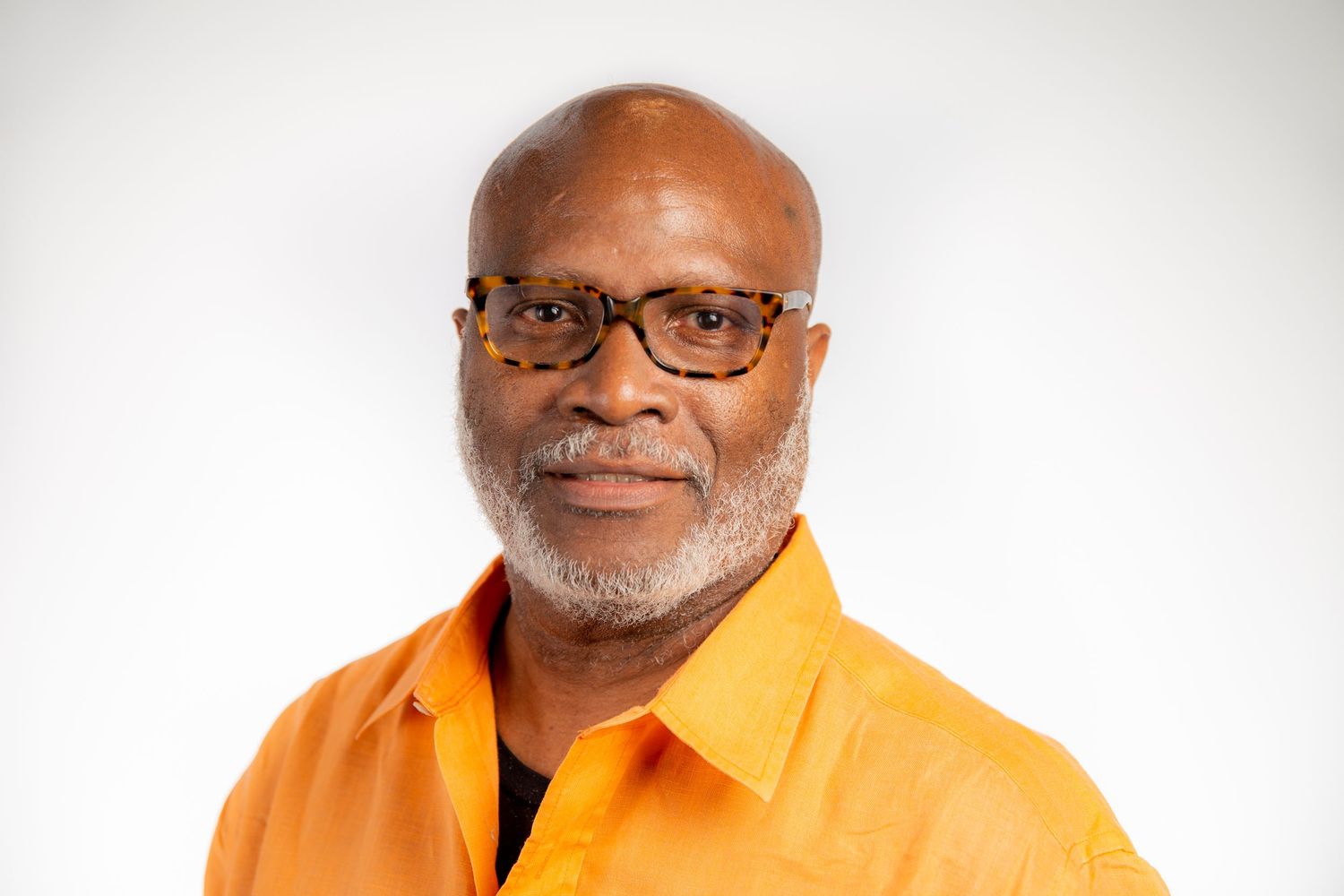 Professional headshot of a smiling bald man with a gray beard wearing an orange shirt on a white background.