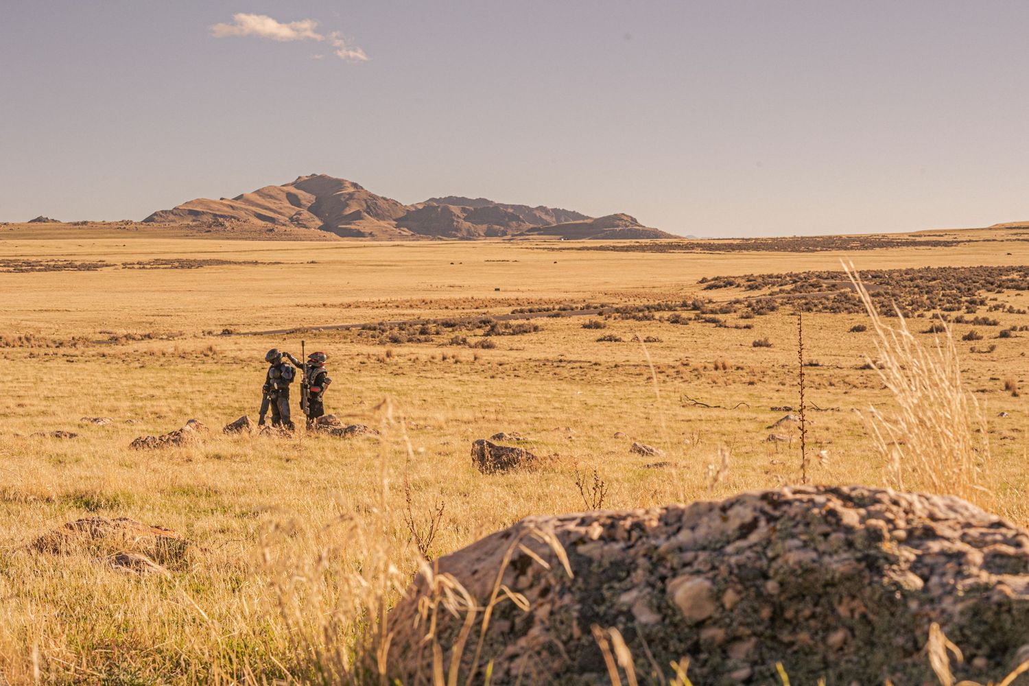 Two cosplayers in dark sci-fi armor stand in vast golden grassland with mountain backdrop.