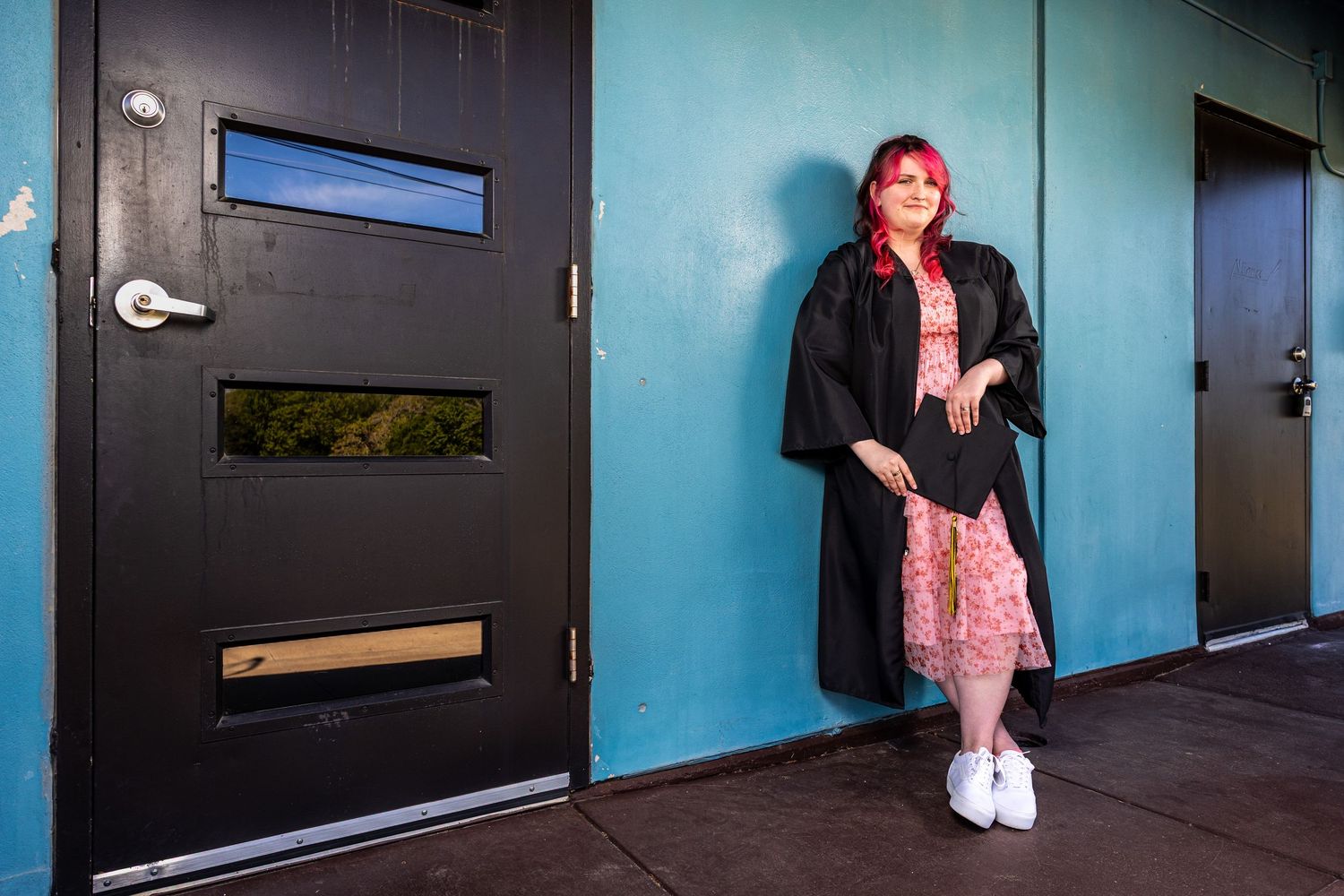 A graduate with pink hair leans against a teal wall beside a black door wearing a black gown.