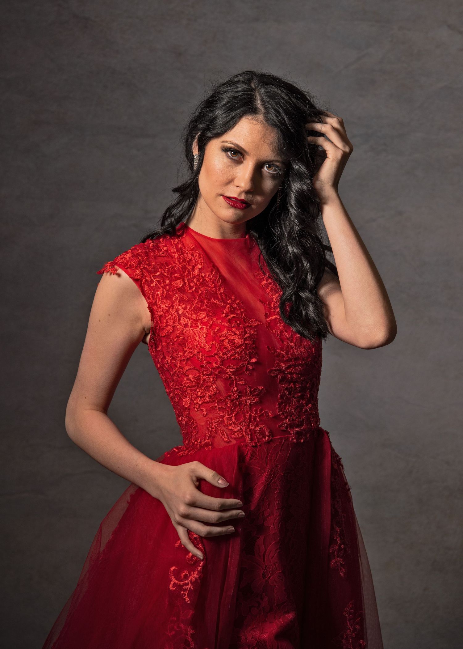 Woman in elegant red lace gown posing gracefully against gray studio backdrop.