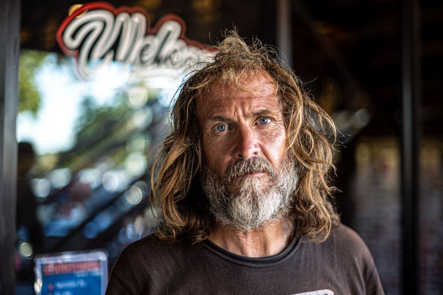 Rugged man with long blonde hair and beard stands outside a bar with neon signage.