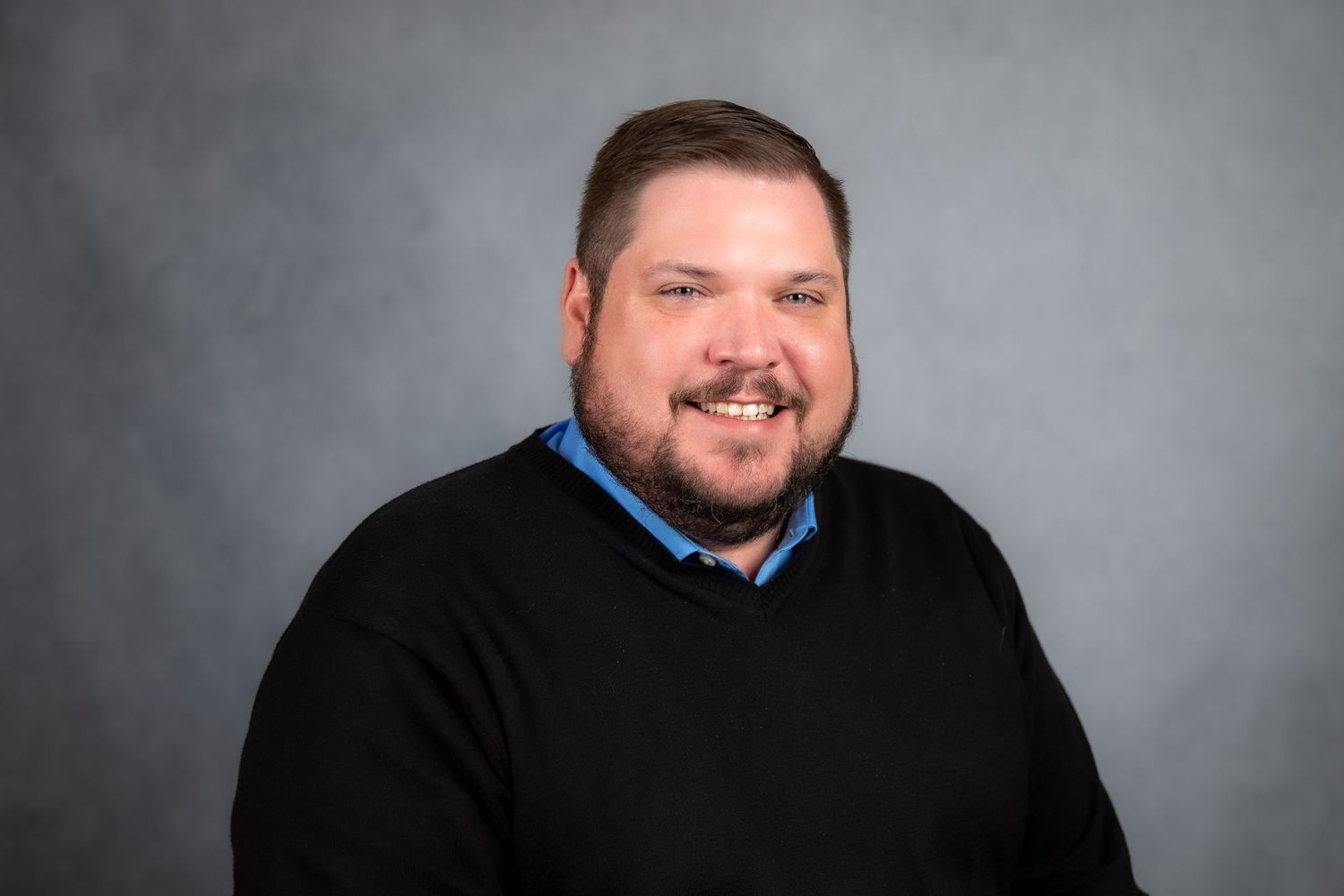Professional headshot of a smiling bearded man in a black sweater and blue collar shirt against a gray background.