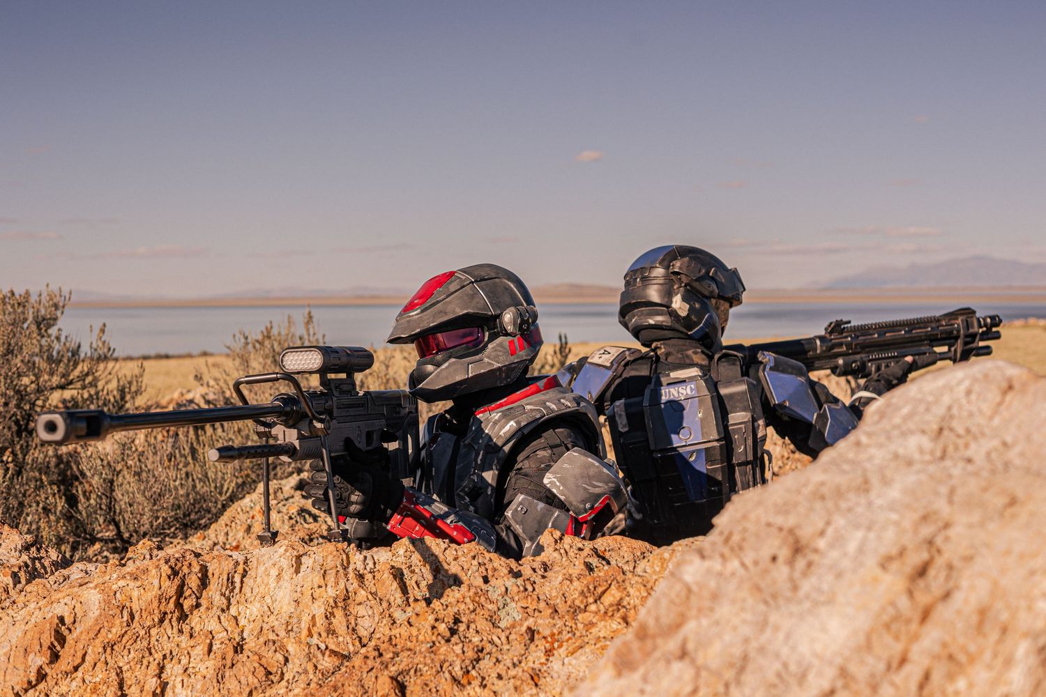 Two airsoft players in tactical armor helmets aiming rifles behind rocky cover near lake.