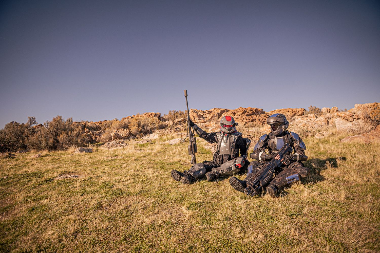 Two Halo ODST cosplayers in full armor rest on grassy hillside holding prop weapons.