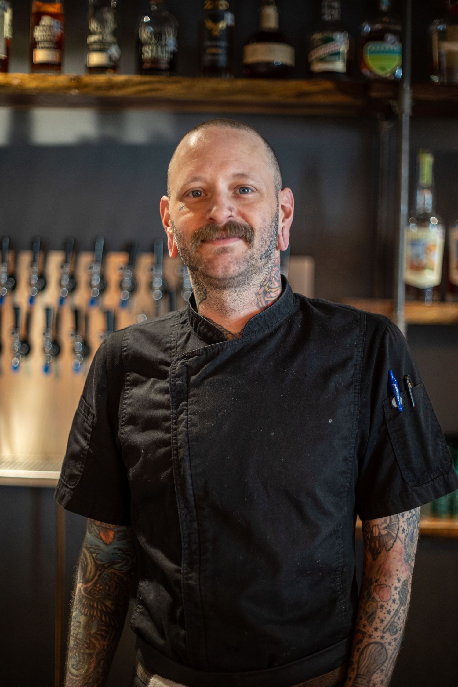 Bald male chef in black jacket with tattoos smiling in front of a bar with beer taps and bottles.