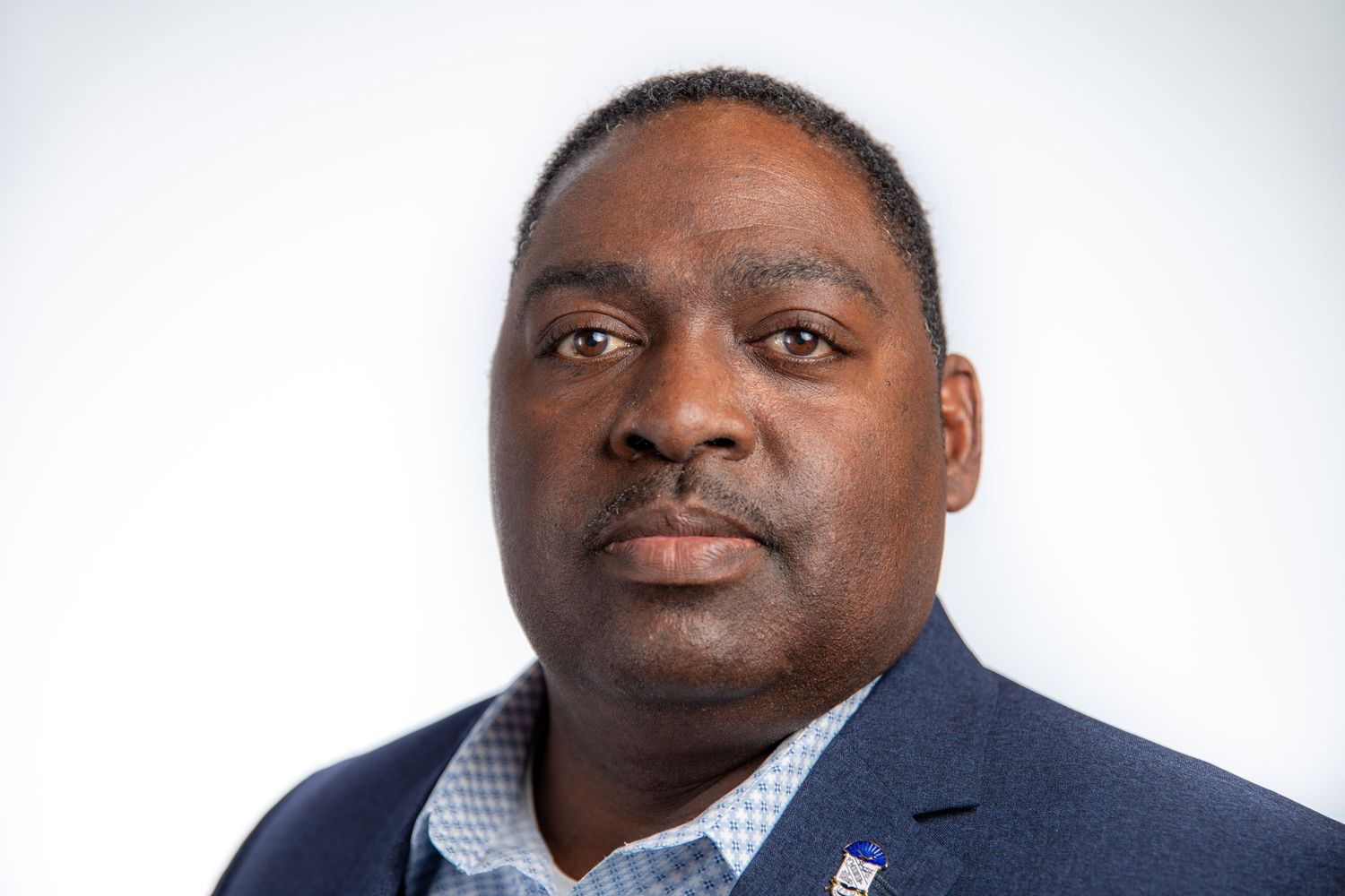Professional Black man in navy blazer with badge posing against a white studio background.