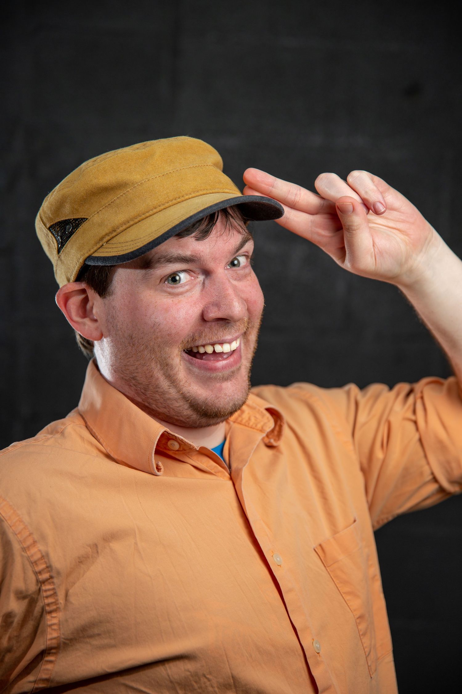 Smiling man in orange shirt tipping a tan cap against a dark background.