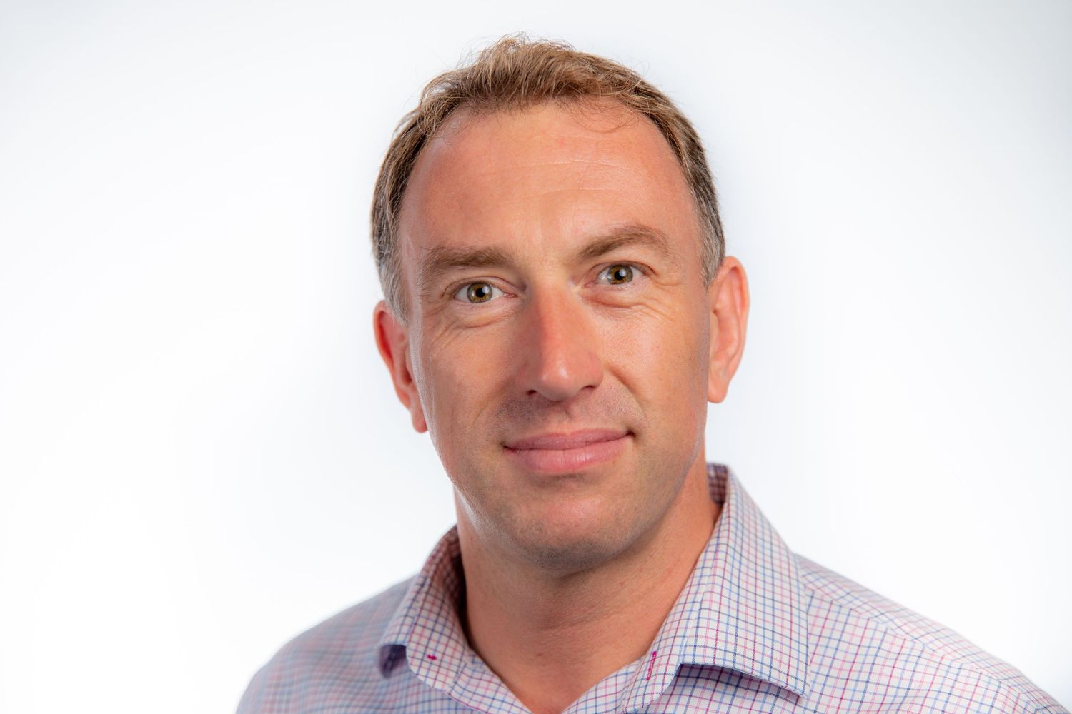 Professional headshot of smiling blonde man in pink checkered shirt on white background.